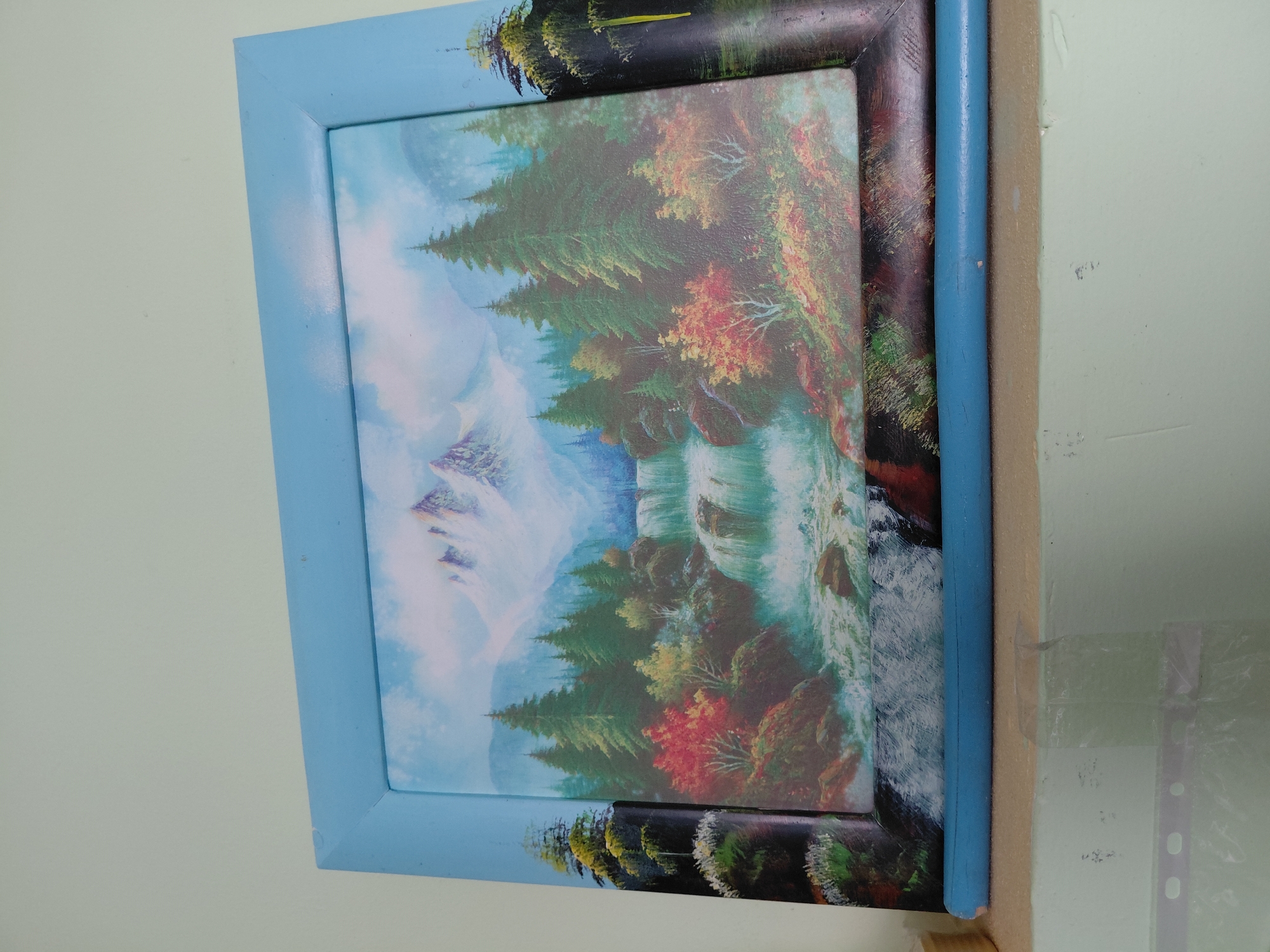 A framed landscape artwork is depicted, positioned against a light-colored wall, likely indoors. The image is oriented vertically, but the artwork itself is in a horizontal (landscape) orientation.
The artwork showcases a vibrant natural scene, dominated by a majestic waterfall cascading down rocky formations on the right side. Dense evergreen trees with sharp, pointed foliage populate the mid-ground and foreground, interspersed with deciduous trees displaying striking autumn colors of red, orange, and yellow. In the background, on the left side, hazy mountains (possibly snow-capped or shrouded in mist) rise under a pale blue sky with soft white clouds. The water at the base of the waterfall appears turbulent and frothy.
The artwork is housed within a light blue, simple wooden frame with mitered corners. This outer frame shows minor scuffs and marks. Inside this frame, the painting appears to have an additional, darker inner border, possibly part of the canvas or a deeper frame, which has some painted elements extending onto it, particularly at the top and bottom edges, mimicking foliage.
To the right of the framed artwork, a light blue cylindrical object, resembling a colored pencil, is visible, tucked vertically between the frame and the wall. Behind it, a beige section of the wall or furniture is discernible, with clear adhesive tape and part of a clear plastic document protector (sleeve with punched holes) partially visible on the bottom right.
The scene in the painting evokes a sense of natural beauty and tranquility, under what appears to be daytime conditions. There is no visible text in the image.