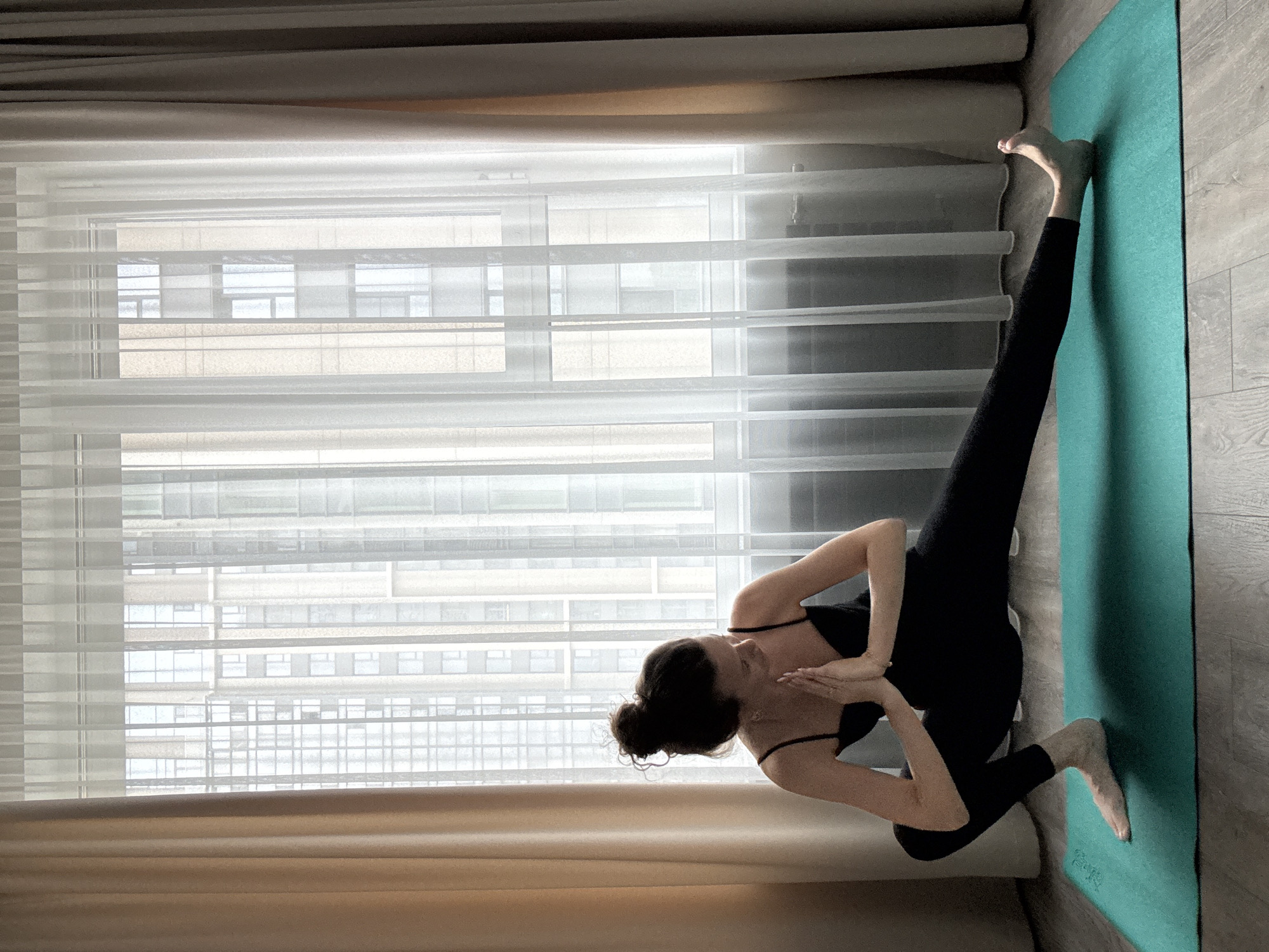 A woman in black athletic wear performs a yoga pose on a teal mat in front of a window with sheer white curtains and beige drapes. The woman is positioned with her left leg bent and her right leg extended outwards, with her hands pressed together at chest level in a prayer position. Behind her, a modern building with many windows is visible through the translucent curtains. The floor appears to be made of wood-like planks.