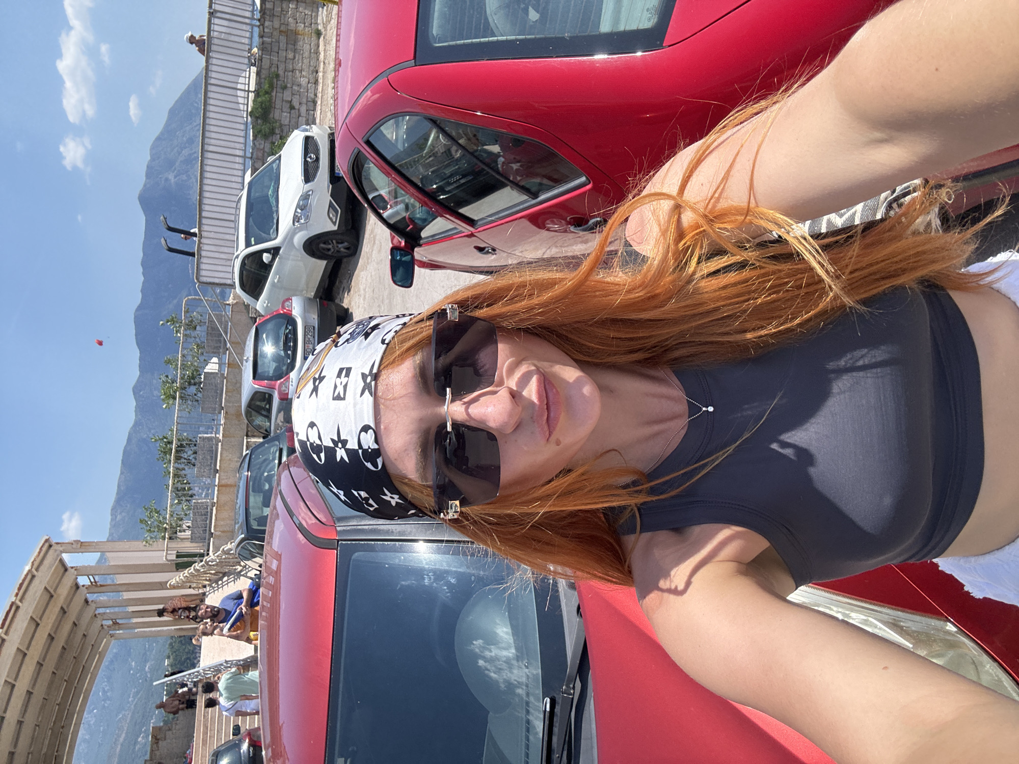 A woman with long, red hair and sunglasses is taking a selfie in front of a red car. She is wearing a dark tank top and a bandana with a black and white pattern. In the background, there are other cars parked, as well as people walking and standing near a staircase and a wall. Mountains are visible in the distance under a clear blue sky with scattered clouds. The location appears to be a parking area near a coastal town, likely Budva, Montenegro, given the mountainous backdrop and the presence of vehicles suggesting a public space.