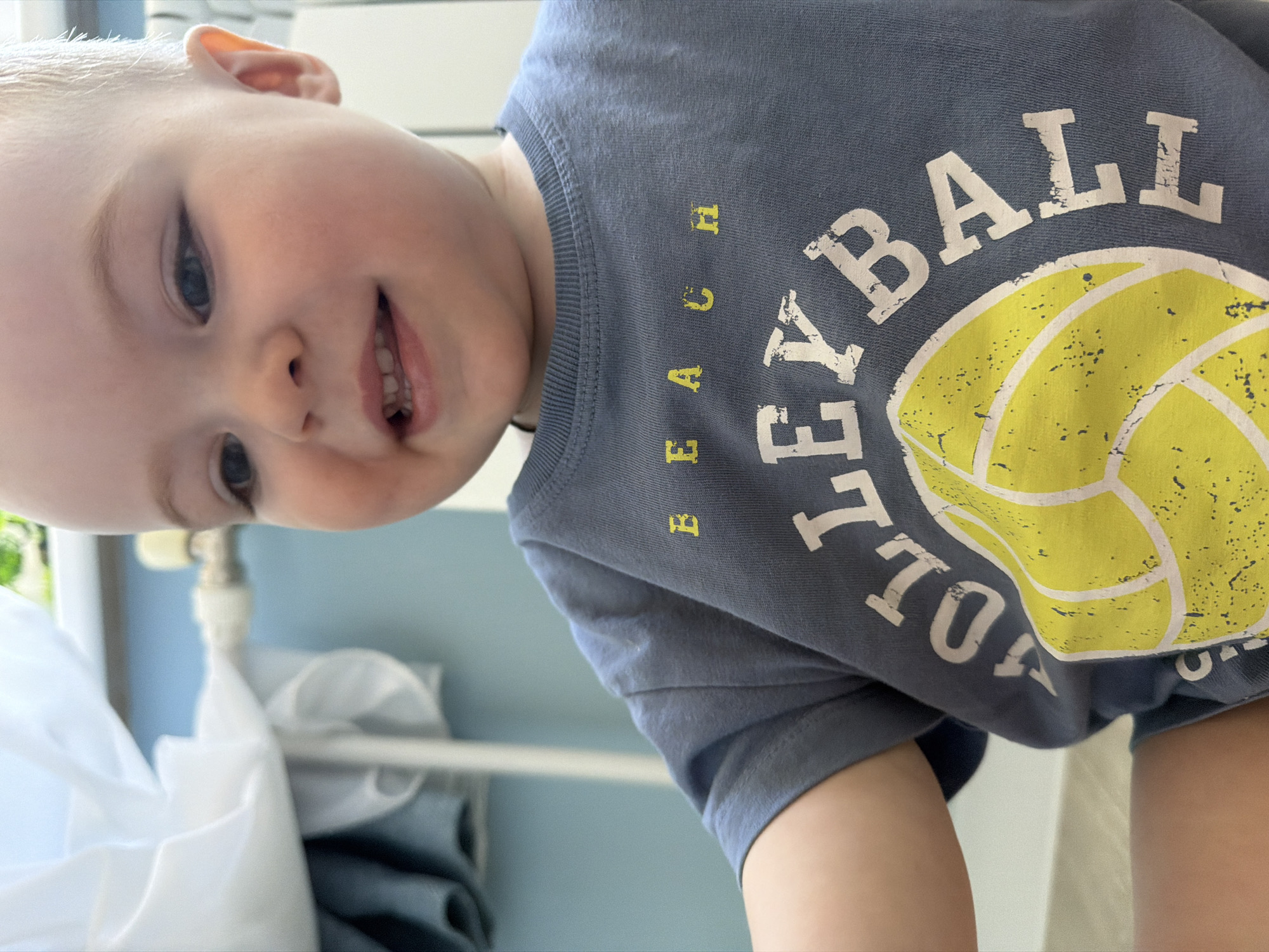 A young child with blond hair and blue eyes is smiling. The child is wearing a gray t-shirt with the word "Volleyball" and a yellow volleyball image on it. The word "Beach" is printed above "Volleyball". The child is indoors and the location is unknown. It is not possible to identify if the location is in Votkinsk, Russia.