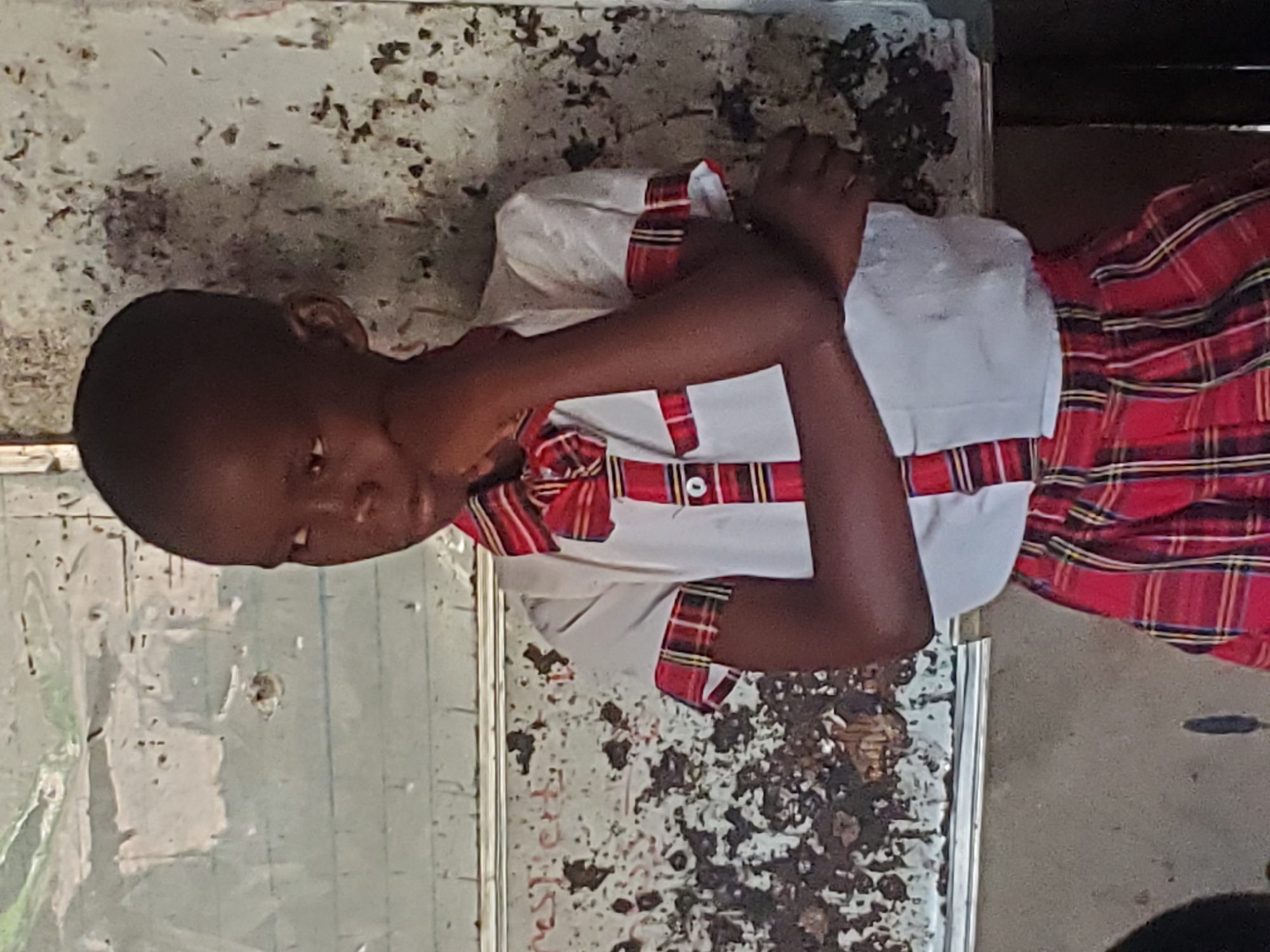 A young person with dark skin, clad in a white shirt with red and white plaid trim and a red plaid skirt, sits indoors with their arms crossed. The background shows a dirty, possibly damaged wall. The location is Unknown, Unknown.