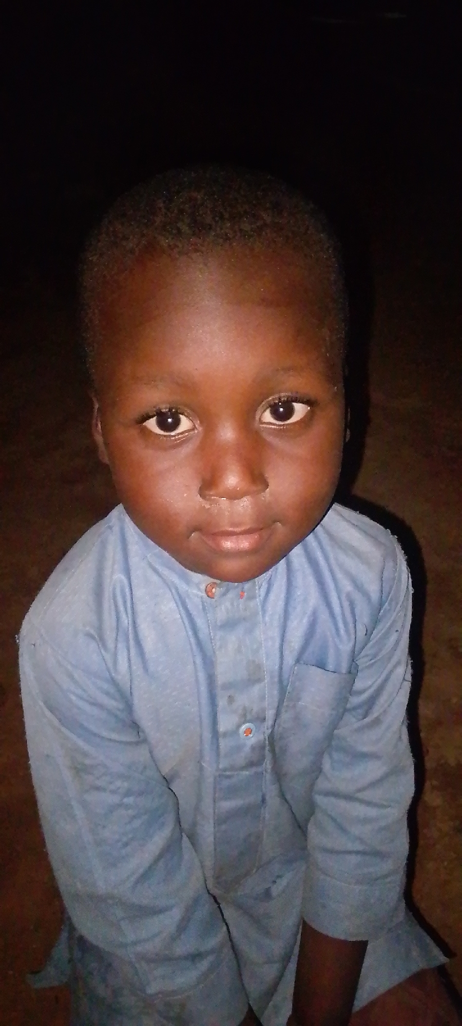 A child with dark skin, wearing a light blue, button-down shirt, is looking directly at the camera. The background is dark, indicating possible low lighting conditions. Details about the specific location in Jos, Nigeria, are not discernible from the image.