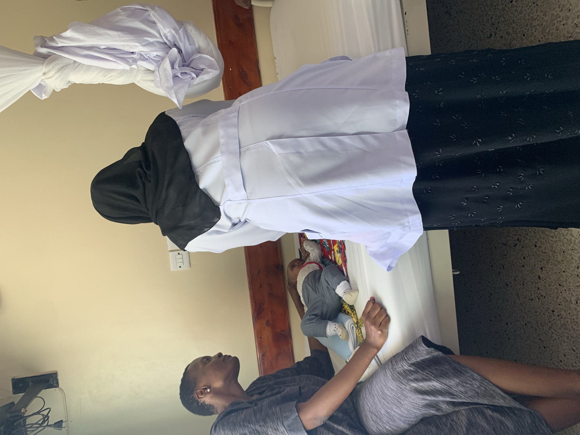 A woman in a black hijab and white medical coat stands over a person lying on a bed. A baby lies on the bed near the person. Another woman sits near the bed. The setting appears to be an indoor location. The city is Dar es-Salaam, Tanzania.