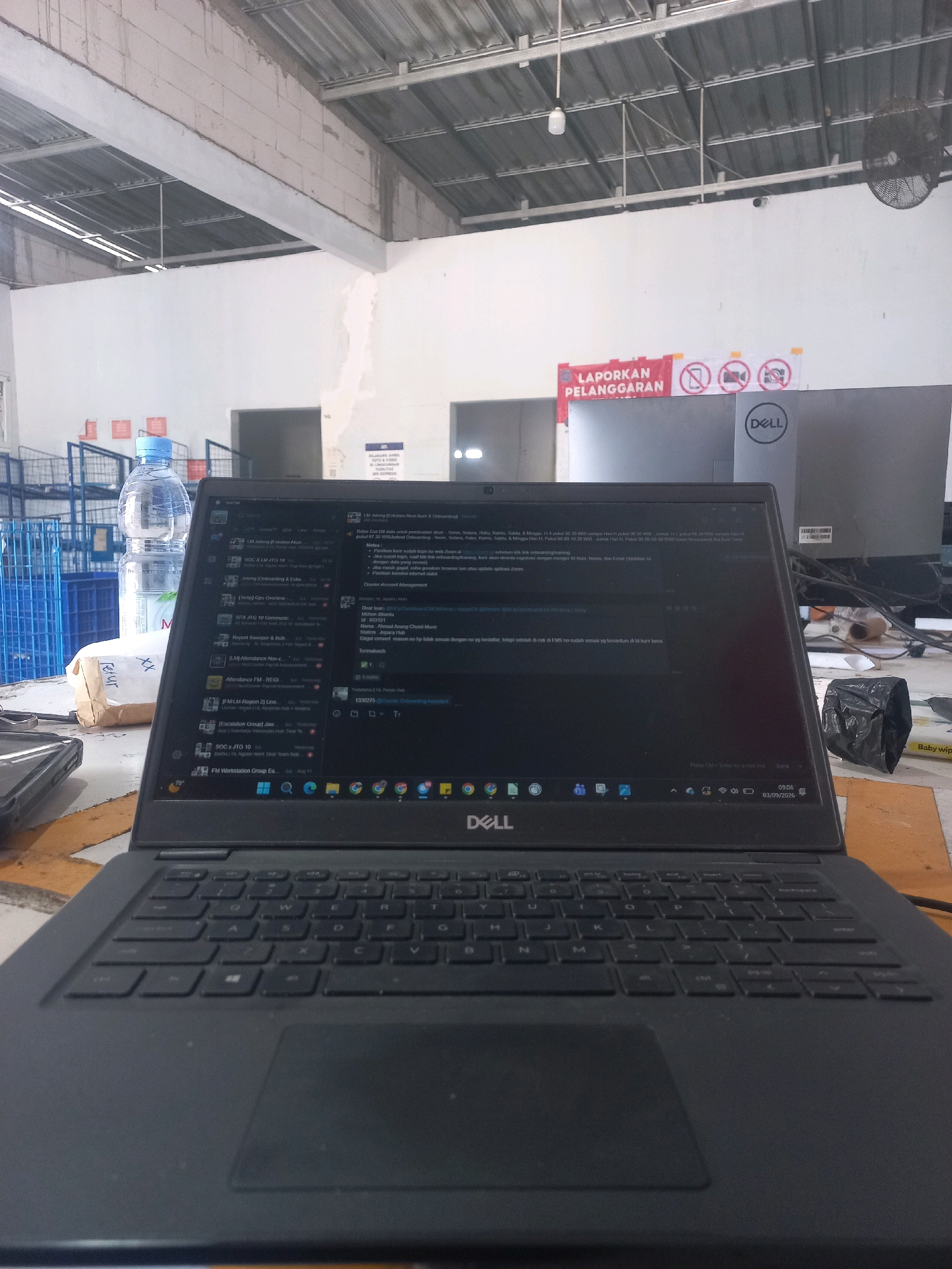 A Dell laptop is open on a table in what appears to be a warehouse or office space in Baturetno, Indonesia. The laptop screen displays an email inbox. In the background, there are shelves filled with blue plastic crates, a water bottle, and a sign that reads "LAPORKAN PELANGGARAN," which translates to "REPORT VIOLATIONS." Other signs with crossed-out camera and phone icons are also visible, suggesting restrictions on photography. The environment is brightly lit by overhead fluorescent lights. The date on the laptop screen shows "03/09/2026".