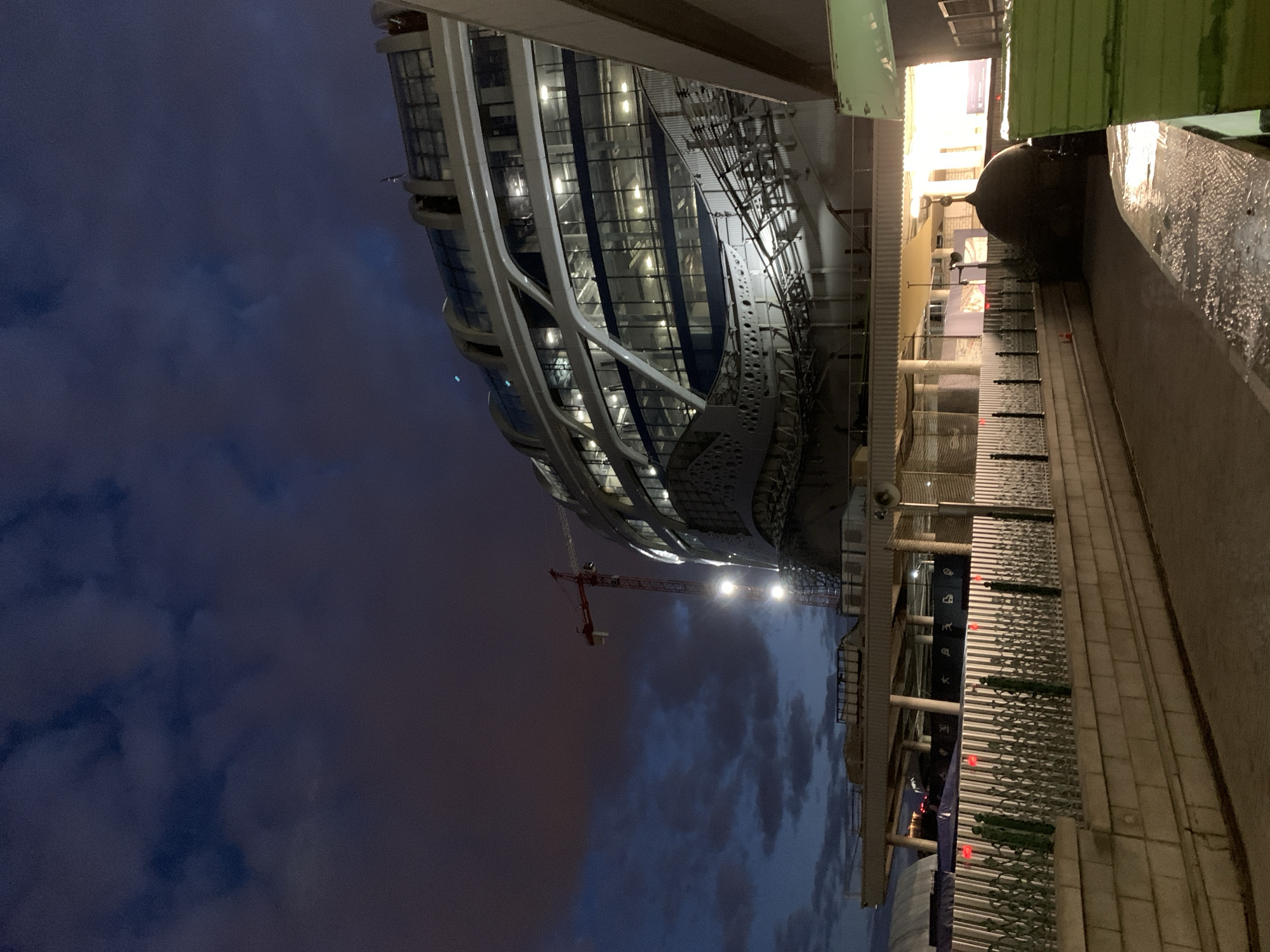 The image depicts a modern building under construction in the city of Unknown, Unknown. The building features a unique design with curved lines and extensive glass windows. A construction crane is visible against a cloudy sky, illuminated by artificial lights. Adjacent to the building, a sidewalk runs along a wall with decorative metalwork and signage. The scene is captured at dusk or night, with the sky transitioning from a deep blue to a lighter hue near the horizon.