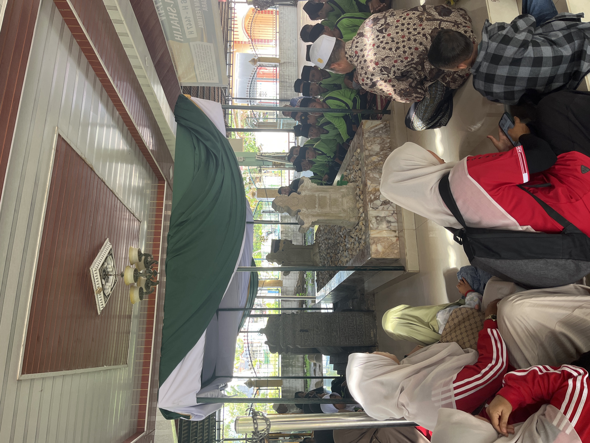 The image shows a group of people gathered at a tomb in Beuringen, Indonesia. The tomb, adorned with carved stone and covered in pebbles, is central to the scene. Several individuals, including women wearing hijabs and men in traditional attire, are seated or kneeling around the tomb. A group of young boys in green shirts and skullcaps are also present, seated attentively. Overhead, a green and white canopy provides shade, supported by poles. The surrounding environment includes a fence, greenery, and buildings with arched windows. A banner with text in Indonesian is visible on the right. The scene appears to be a religious gathering or a visit to a sacred site.