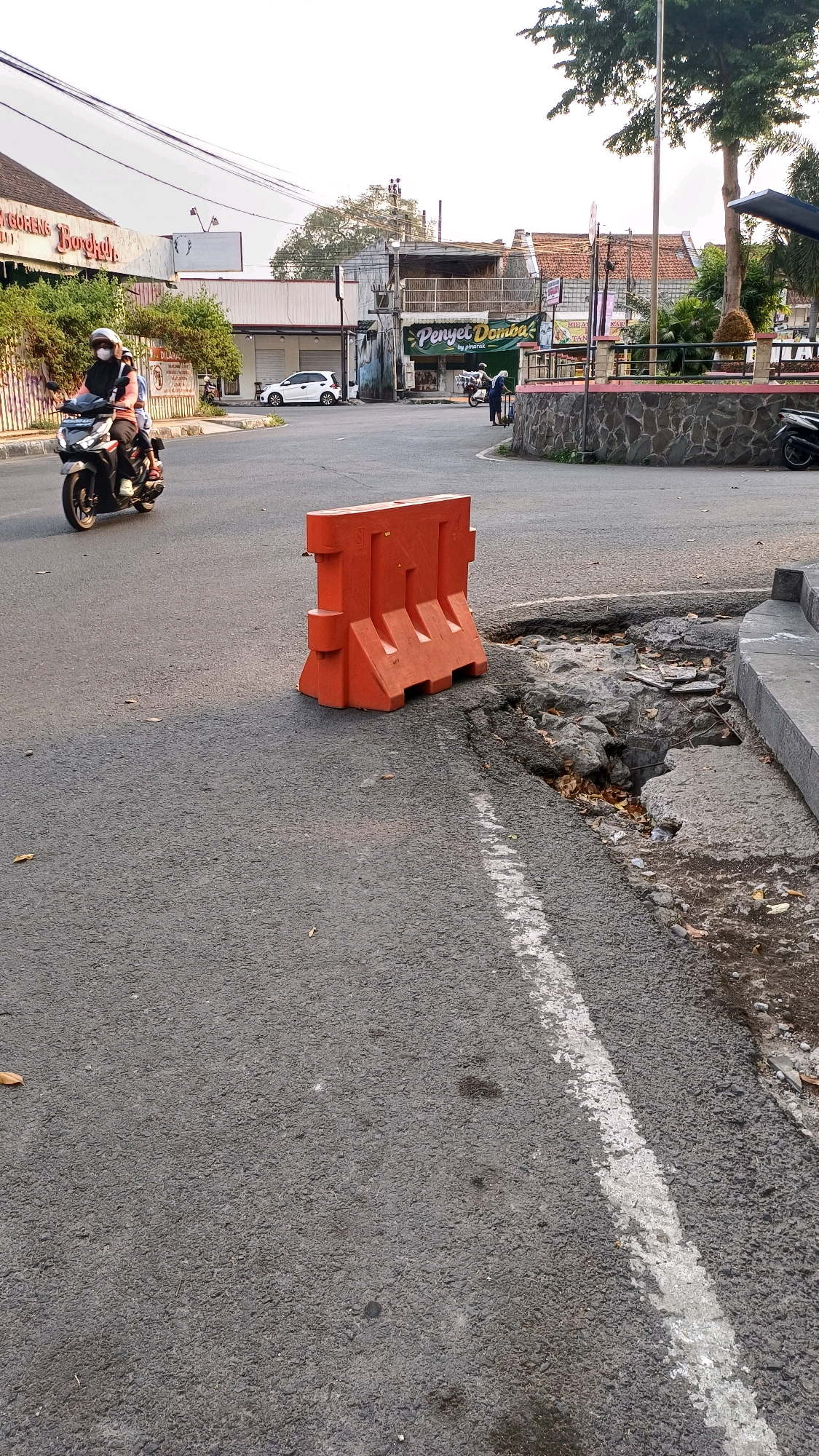 An outdoor, eye-level, medium shot captures a section of road in Klaten, Indonesia, with a focus on a bright orange plastic barrier placed to cordon off a damaged section of pavement. The barrier, a typical traffic control device, stands prominently in the foreground, its color contrasting with the gray asphalt. To the right of the barrier, a significant pothole or sinkhole has formed in the road, revealing exposed rocks and debris. A white dashed line runs along the asphalt, indicating a lane marker. In the middle ground, a white car is parked on the left side of the road. Further back, several buildings line the street, some appearing to be businesses with signs. One sign, partially visible, reads "Penyet Domba." On the left side of the road, a person rides a scooter, wearing a helmet and appearing to be a passenger on the back. A single tree is visible in the background, its branches reaching towards the sky. The overall environment suggests a somewhat urban or suburban setting with evidence of road maintenance issues.