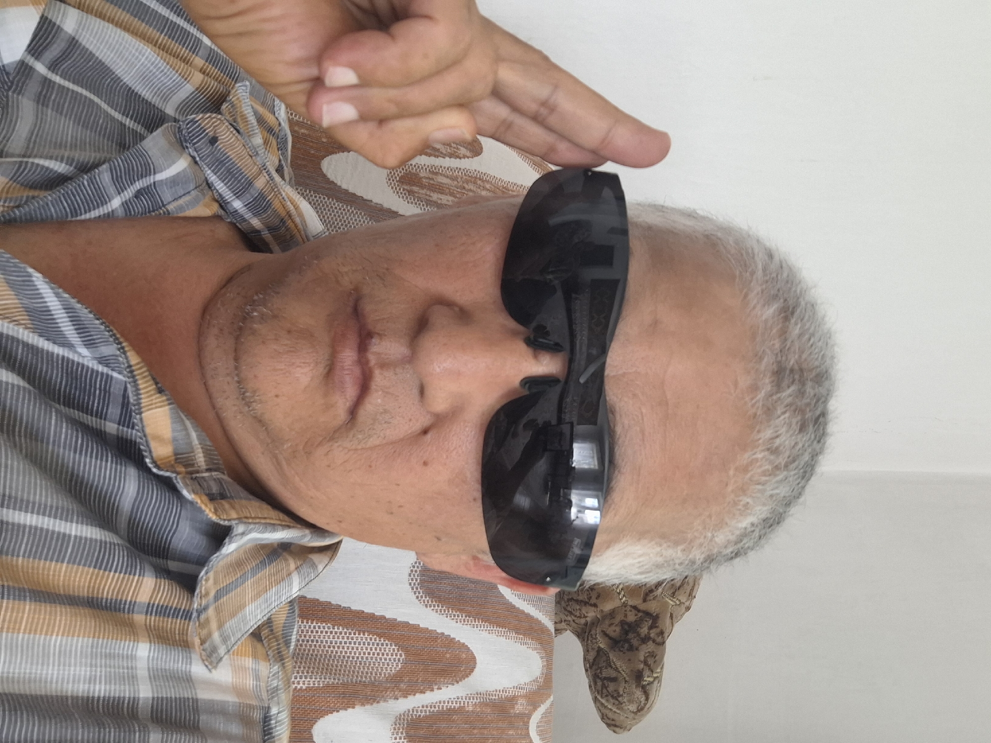 A middle-aged man with graying hair and a plaid shirt is posing for a selfie. He is wearing dark sunglasses and has his right hand raised to the side of his head. He appears to be indoors, with a white wall behind him and a patterned couch visible to the right. There is a decorative object on the couch that resembles a dinosaur or lizard. The lighting suggests it is daytime. The image does not provide any specific information about its location within the city of Almaty, Kazakhstan.