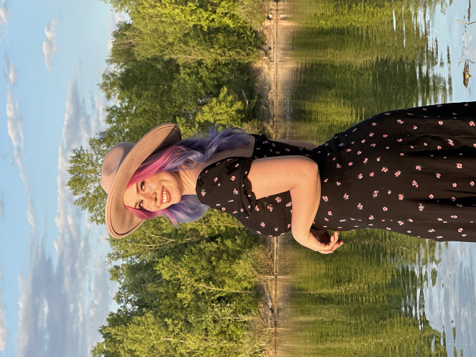 A vibrant portrait captures a young woman outdoors during what appears to be the golden hour, with warm light illuminating the scene. She is seen from the waist up, smiling brightly towards the camera, suggesting a posed photograph. Her hair features striking pink and purple tones, partially covered by a light brown, wide-brimmed hat. She wears a black dress adorned with small pink floral patterns, and subtle glitter or face gems are visible on her left cheekbone.
The background shows a serene natural environment. Behind her is a body of water, likely a lake or pond, reflecting the surrounding trees and sky. Lush green trees line the far bank, and above them, a clear sky with scattered white clouds indicates fair weather. The woman's joyful expression contributes to the overall happy and tranquil mood of the image. The user has specified the location context as the City of Zagreb, Croatia. There is no visible text in the media file.