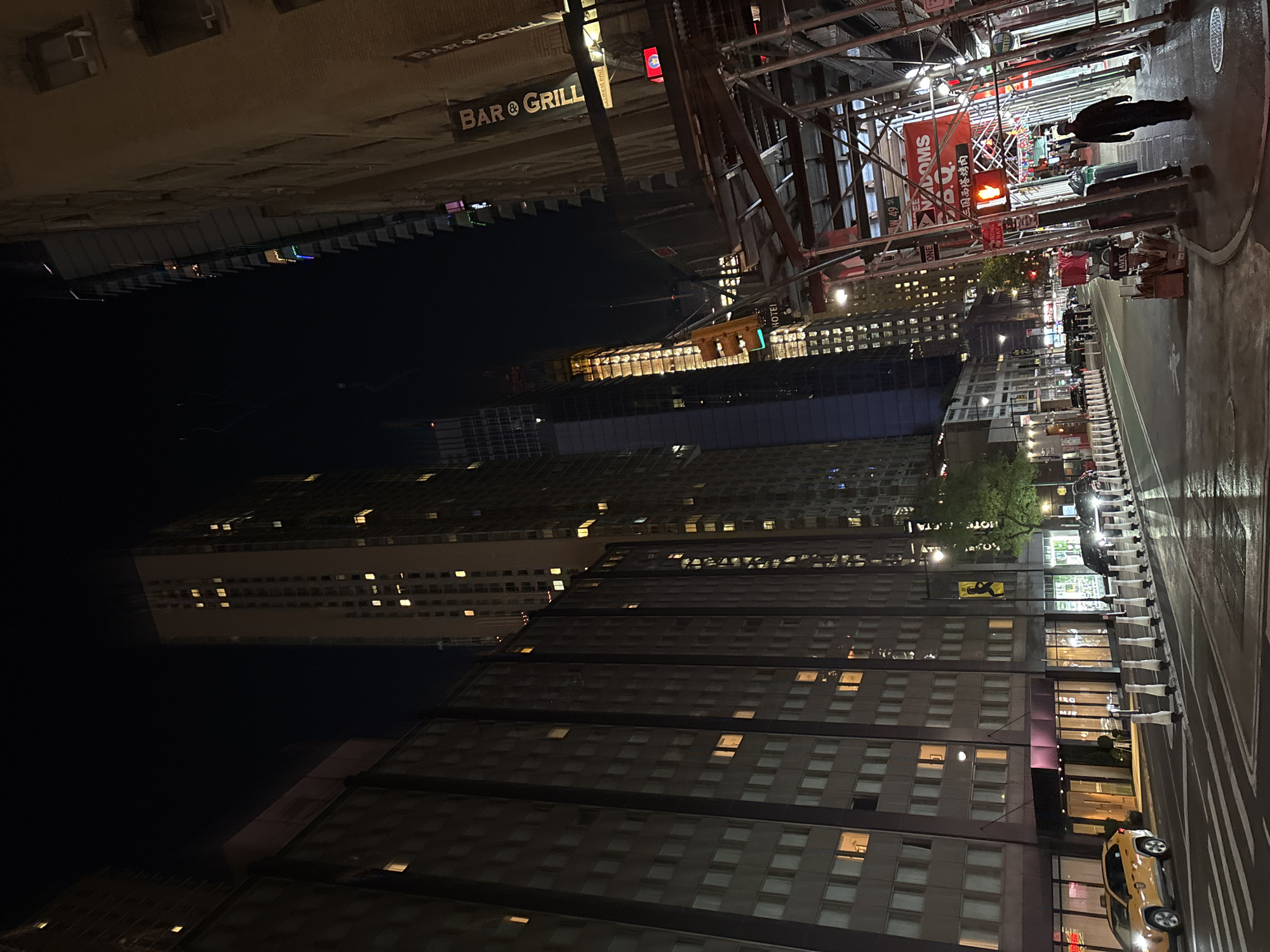 The image captures a nighttime street scene in a dense urban environment, characteristic of New York City. The perspective appears to be slightly elevated, looking down a street lined with towering buildings.
The time of day is night, with the sky completely dark, contrasting sharply with the myriad of illuminated windows in the surrounding high-rise buildings and the bright streetlights. The street surface appears somewhat reflective, suggesting it might be wet or has been recently.
In the mid-ground, a pedestrian, silhouetted in a dark coat, walks on the sidewalk underneath a large scaffolding structure that runs along the right side of the street. A red pedestrian signal displaying a "DON'T WALK" hand symbol is visible near the scaffolding. A row of white street barriers or bollards separates the pedestrian area from the street, where several vehicles, including a distinctive yellow taxi, are present.
Visible text on signs throughout the scene includes "BAR & GRILL" (appearing multiple times), "HOTEL," "W. 49 St," "MADISON S.B.Q.," "ONE WAY," and Chinese characters "三国西塔烤肉" (translating to "Three Kingdoms West Tower BBQ"). The presence of a yellow taxi and the street name "W. 49 St" strongly indicates the location is New York City, United States of America. The scene depicts a lively, artificially lit urban landscape with a mix of commercial and residential buildings.