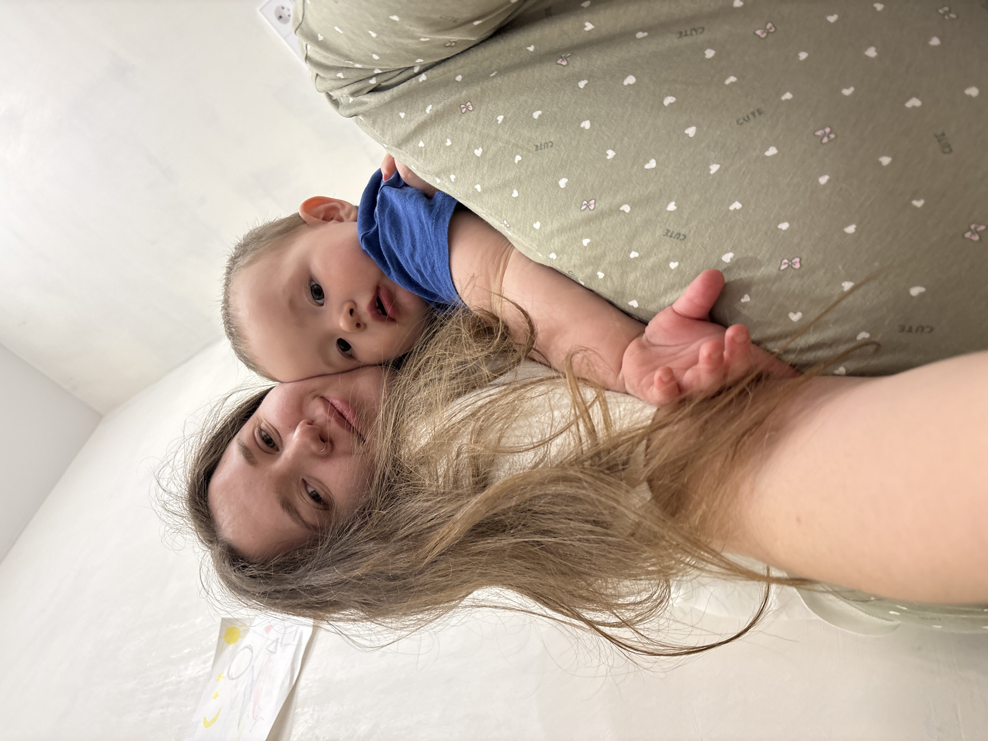 This image depicts two individuals in an indoor domestic setting, located in the city of Izhevsk, Russia.
**People:**
An adult female, positioned on the left side of the frame, is looking directly at the camera with a faint smile. She has long, light brown hair, a fair complexion, and a small piercing on her right nostril. She is wearing a light-colored, possibly white or off-white, top. Her left arm is visible, supporting an infant.
An infant, positioned to the right of the female, is looking towards the camera with a neutral expression and slightly open mouth. The infant has light brown hair and is wearing a blue short-sleeved top.
**Objects:**
The female is also wearing olive-green bottoms (trousers or sleepwear) patterned with small white hearts, pink bow motifs, and the word "CUTE" repeated.
In the background, two pieces of paper with children's drawings are affixed to a light-colored, possibly white, wall. One drawing on the upper left depicts a yellow sun and other abstract shapes. Another drawing, further right, shows a simplified face or abstract design.
**Actions and Interactions:**
The female is holding the infant in a close embrace. Both individuals are oriented towards the camera, suggesting a selfie or close-up photograph.
**Environment:**
The background consists of plain, light-colored walls, which appear to have a slightly textured finish. The lighting is even and originates from an unseen source, likely overhead, illuminating the scene brightly. The overall environment suggests a private residence or a similarly informal indoor space.