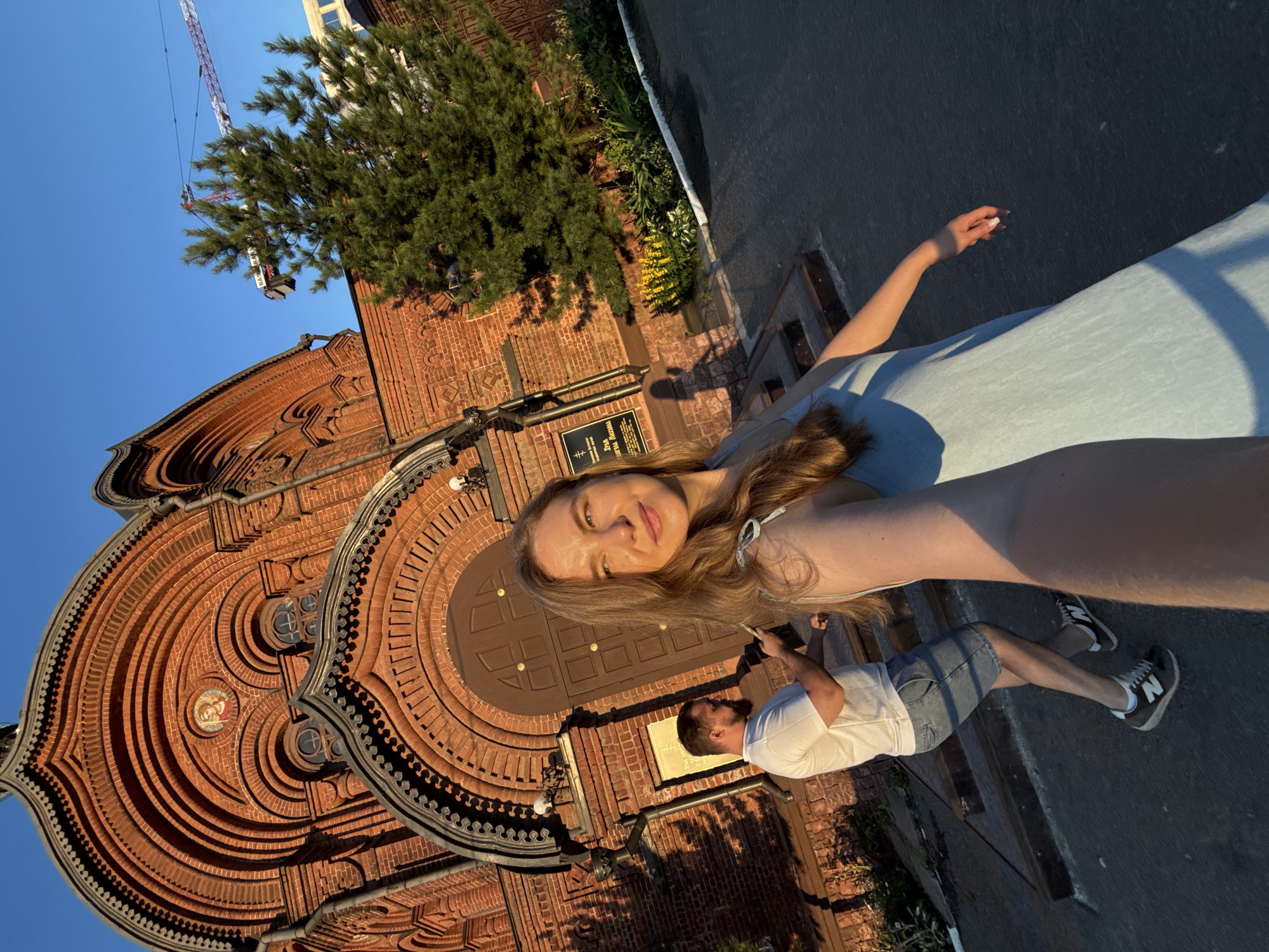 The image shows a woman in a light blue dress taking a selfie in front of a brick building with arched windows and a brown wooden door. A man in a white t-shirt and denim shorts is walking away from the camera in the background, holding a phone. A tree with green foliage stands to the right of the building, and a construction crane is visible in the distance against a clear blue sky. The setting appears to be outdoors, likely on a sunny day, given the shadows and the warm light on the building. The architecture of the building suggests it could be a church or a historical structure. Based on the available information, the building in the image is the Church of St. Nicholas the Wonderworker, located on the embankment of the Klyazma River in the city of Vladimir, Russia.