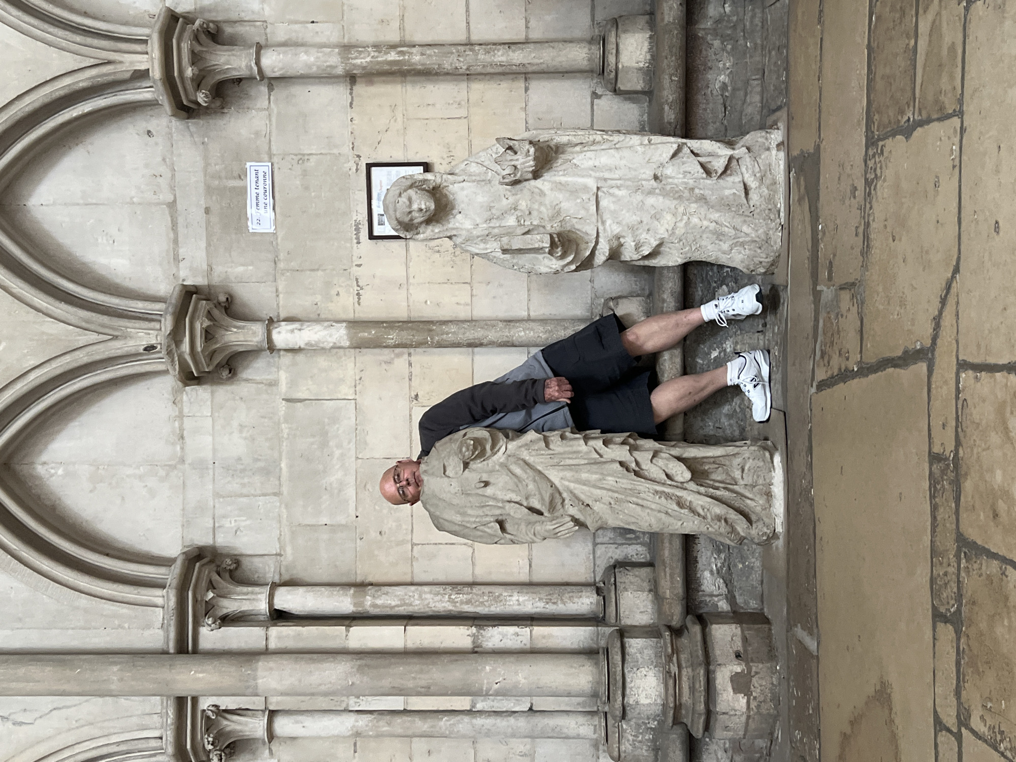 The image captures a man posing between two ancient stone statues inside a grand, historic building, likely a cathedral or church in Rouen, France. The man, who is bald and wears glasses, a grey long-sleeved shirt, black shorts, and white sneakers, leans casually against the statue on the left, looking at the camera with a neutral expression.
Both statues are made of weathered, light-colored stone and depict robed figures standing on pedestals, typical of religious or historical sculptures. The statue to the man's right is specifically identified by a small white sign on the wall behind it, which reads "22. femme tenant une couronne" (22. woman holding a crown). This indicates the statue depicts a woman holding a crown.
The setting features light-toned, aged stone walls constructed from large blocks. Intricate architectural details such as decorative pilasters, cornices, and arched relief patterns are visible, reinforcing the impression of a significant historical structure. The lighting is soft and even, suggesting an interior space that is well-lit, possibly by natural light filtering in or ambient artificial light. The scene depicts a tourist engaging with the historical art and architecture of the location.