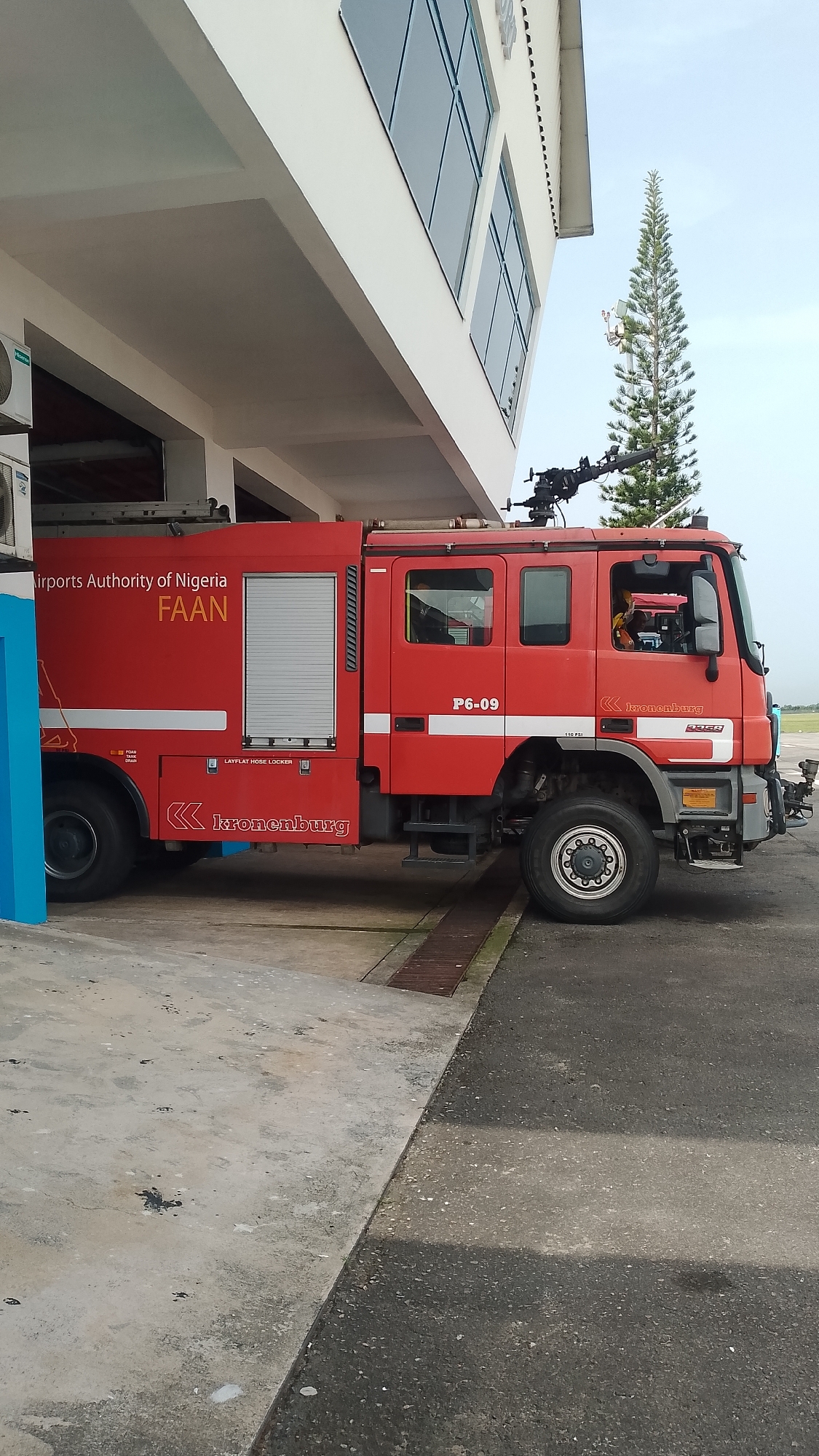 A red Kronenburg fire truck, identified as "P6-09" and operated by the Airports Authority of Nigeria (FAAN), is parked partially under the overhang of a two-story white building with large windows. The vehicle features a turret-mounted nozzle. The setting includes a paved area transitioning from smooth concrete to asphalt and a drain grate. A tall, coniferous tree is visible in the background. The location is identified as Orerokpe, Nigeria. There are no people visibly outside of the vehicle.
