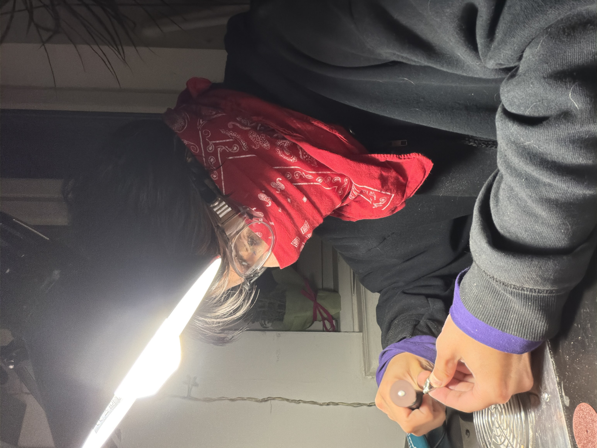 A person wearing glasses and a red bandana is hunched over, working on jewelry with a handheld tool under bright light. The person is wearing a black zippered hoodie with purple cuffs. The workspace includes tools and materials scattered on a surface. The background is indistinct, suggesting an indoor location. The city of Sofia, Bulgaria cannot be determined from the image.