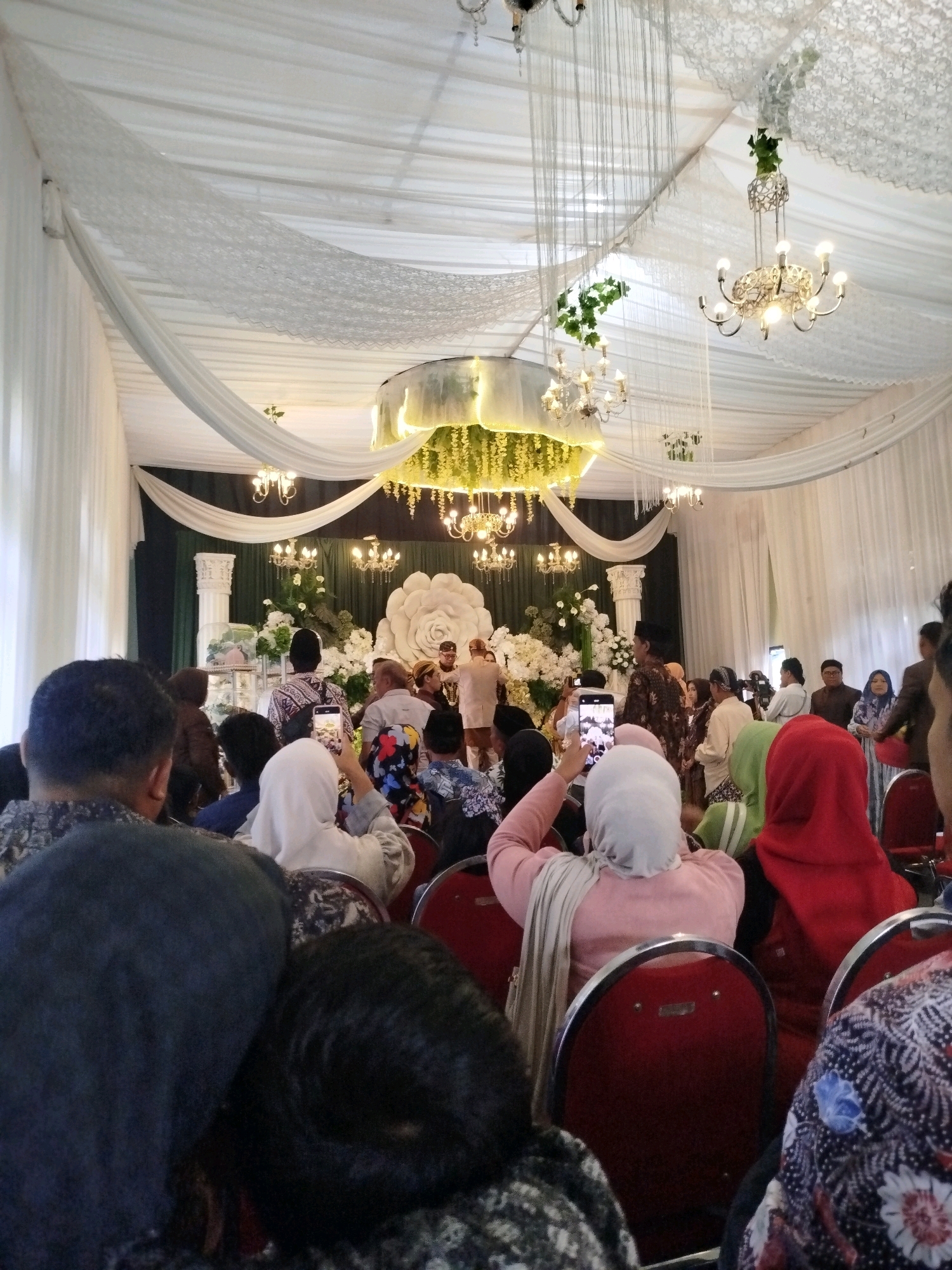 The image captures an indoor wedding reception, likely in Malang, Indonesia, judging by the architectural style and the attire of the attendees. The venue is elaborately decorated with white draped fabric covering the ceiling and walls, creating a festive and elegant ambiance. Several chandeliers hang from the ceiling, some adorned with greenery, casting a warm light on the scene.
In the background, a stage area is set up with floral arrangements, including a large, white floral backdrop, and two columns that frame the central area. A wedding ceremony or reception event is taking place on the stage, with several individuals dressed in traditional attire. Some attendees are actively participating in the event, while others are seated in rows of red chairs, observing. Many people in the audience are holding up their phones to capture photos or videos of the ceremony.
The foreground of the image is occupied by the backs of several guests, indicating the photographer is among the attendees. The guests are dressed in a variety of formal and semi-formal clothing, with some women wearing headscarves. The overall impression is of a joyous celebration, filled with family and friends gathered to witness and document the wedding. The lighting is generally soft, with some spotlights directed towards the stage area. The image appears to have been taken with a mobile phone camera, as suggested by the framing and resolution.