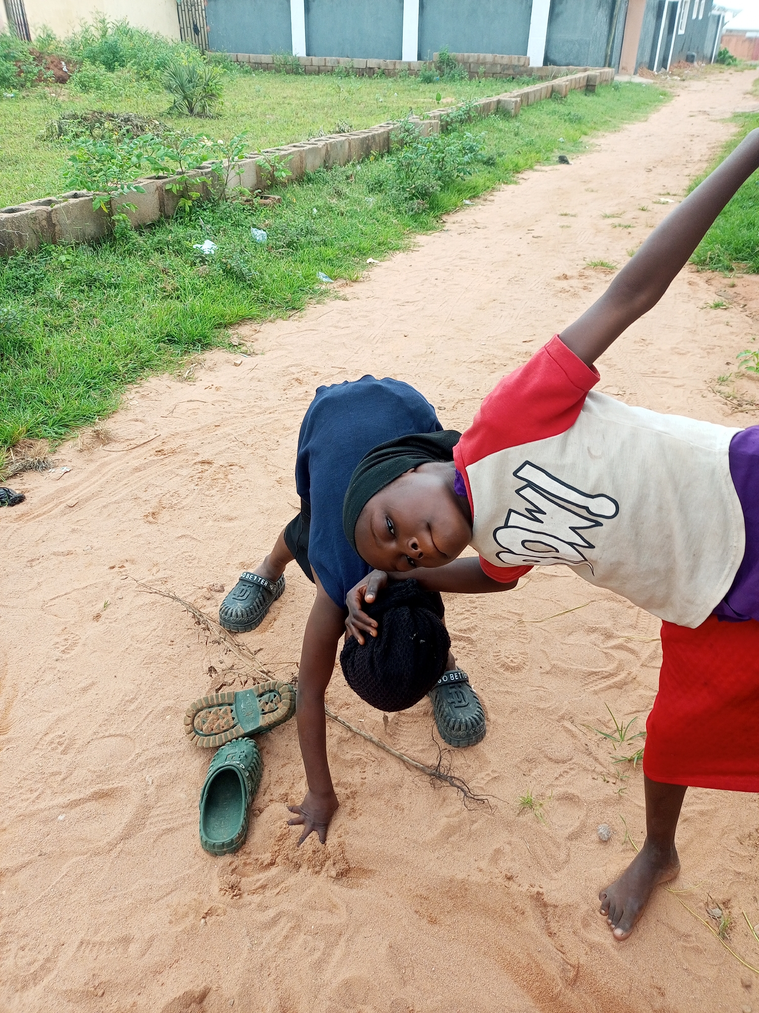 Two children are interacting on a sandy path in front of some buildings in Tempure, Nigeria. The child on the left is wearing a dark blue shirt and black head covering, with black shorts. They are on their hands and knees, looking towards the camera. To their left, a pair of green sandals and the sole of a shoe are lying on the sand. The child on the right is wearing a red and white t-shirt with black lettering and a red skirt. They are leaning backwards with their right arm extended upwards, also looking towards the camera. The environment consists of a dirt path, grass, and low-rise buildings in the background.
