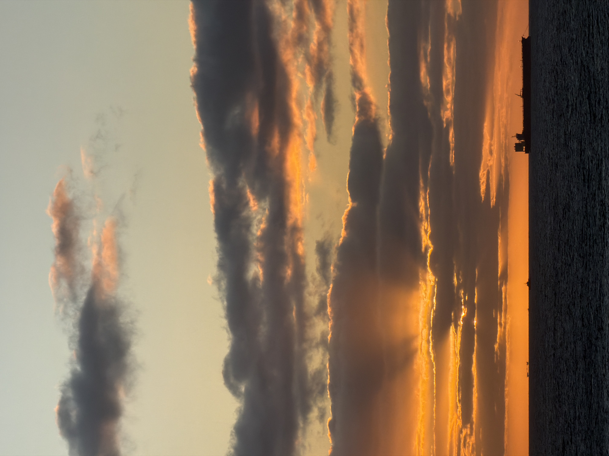 The image shows a sunset over water, likely a sea or large lake. The sky is filled with dramatic clouds, with some appearing dark gray and others lit up by the setting sun, creating an orange or golden glow. The horizon line includes the dark silhouette of what appears to be a ship or other maritime structure. The water is dark and textured, suggesting some waves or ripples. The location and city are unknown from this image.