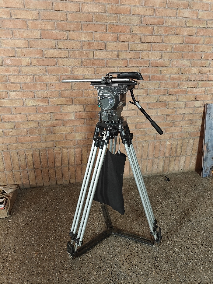 A professional-grade video camera tripod stands centered in the frame against a red-brown brick wall. The tripod features three silver-colored telescopic legs with black locking mechanisms, supported by a black ground spreader connecting the feet. A black fabric bag hangs from the tripod's mid-section. Mounted atop the legs is a dark grey fluid head, equipped with various control knobs, a visible green indicator light, and a black pan bar extending to the right. A silver-colored quick-release plate extends horizontally to the left from the fluid head. The floor is a speckled grey-brown concrete surface. In the far left foreground, a partially visible white object with red markings rests on the ground. To the far right, a dark, rectangular object, possibly a framed picture or board with abstract markings, leans vertically against the brick wall. No human subjects or discernible actions are present. The precise location within Kalyan-Dombivli, India, is not discernable from the visual information provided.