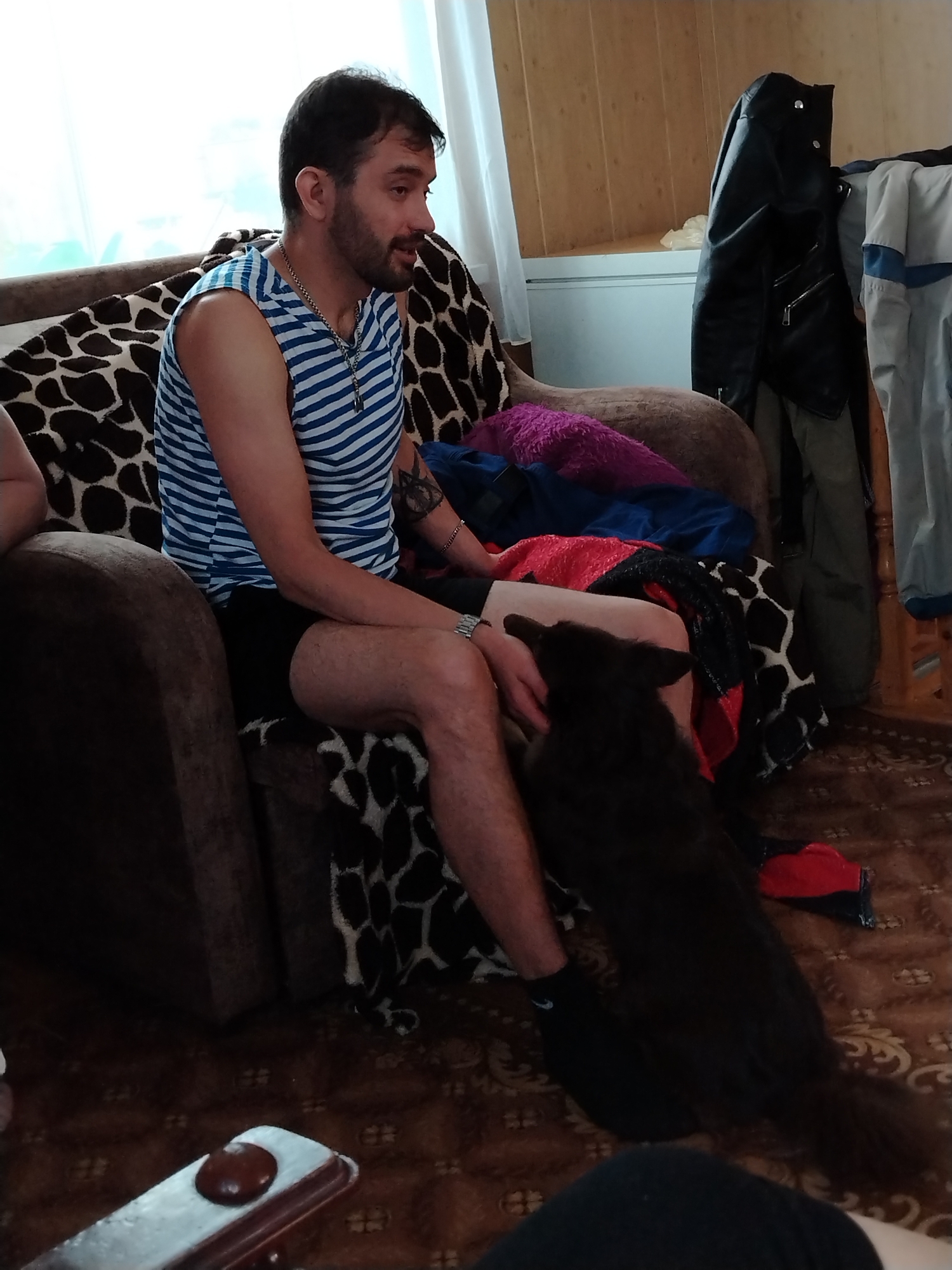 The image shows a man sitting in an armchair in what appears to be a living room. The man is wearing a blue and white striped sleeveless shirt and black shorts. He has a beard and is wearing a necklace. He is seated in a brown armchair with a black and white patterned throw blanket. A black dog is standing on its hind legs in front of the man, its head near his lap. The floor is carpeted with a brown patterned rug. Behind the armchair, there is a wall with what looks like a wardrobe or storage unit, on which a black leather jacket is hanging. A window with white curtains is visible in the background. The scene is in the city of Большая Уса, Russia.