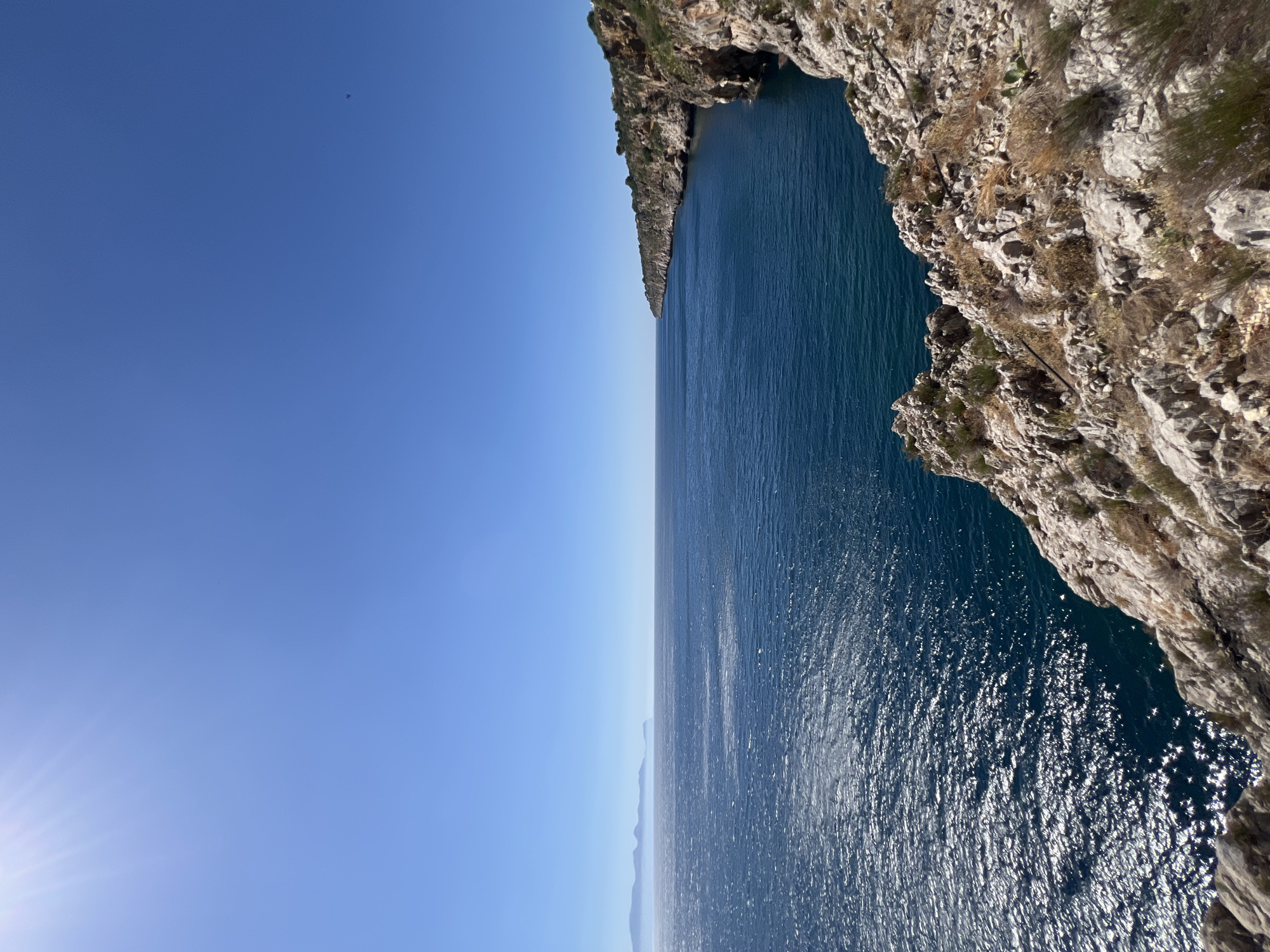 The image depicts a panoramic view of the coastline near Terrasini, Italy. The scene is dominated by the vast expanse of the Mediterranean Sea, which is a deep, vibrant blue and reflects the bright sunlight, creating a shimmering effect on its surface. To the right, rugged cliffs rise steeply from the water, their rocky faces displaying shades of brown and gray, interspersed with sparse green vegetation. The top of the cliffs is covered with more foliage, with some structures faintly visible, suggesting human habitation. In the distance, a hazy, mountainous coastline stretches across the horizon, partially obscured by atmospheric conditions. The sky above is a clear, bright blue, with the sun, a bright white orb, positioned in the upper left corner, casting sunbeams across the sky. The foreground is characterized by rough, rocky terrain covered with dry grass and scrub. There are no people or animals visible in the image, and no specific actions or interactions are taking place. The overall impression is one of a serene and natural coastal landscape.