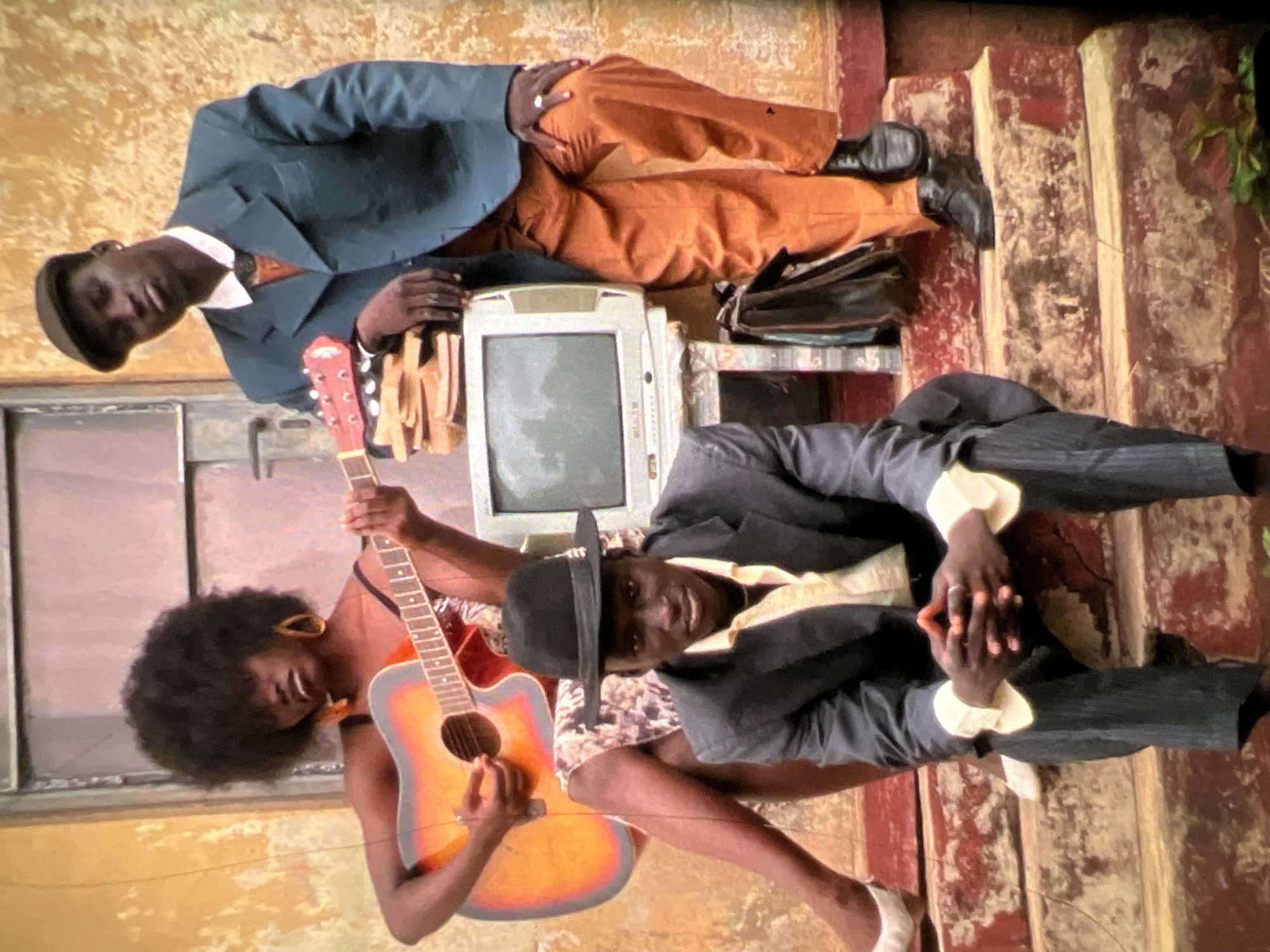 The image depicts three individuals on weathered steps outside a building with a textured, ochre-colored wall. In the foreground, a person sits with hands clasped, wearing a dark suit, a light-colored shirt, and a black fedora. To the left, a person with a prominent afro, wearing a patterned dress and large hoop earrings, plays an acoustic guitar. To the right, a person in a teal suit, brown pants, and a cap leans against a stack of books atop an old television. A briefcase rests on the steps below them. The scene appears to be an outdoor urban setting in Lagos, Nigeria, possibly a residential area or an older part of the city, given the worn appearance of the steps and building.