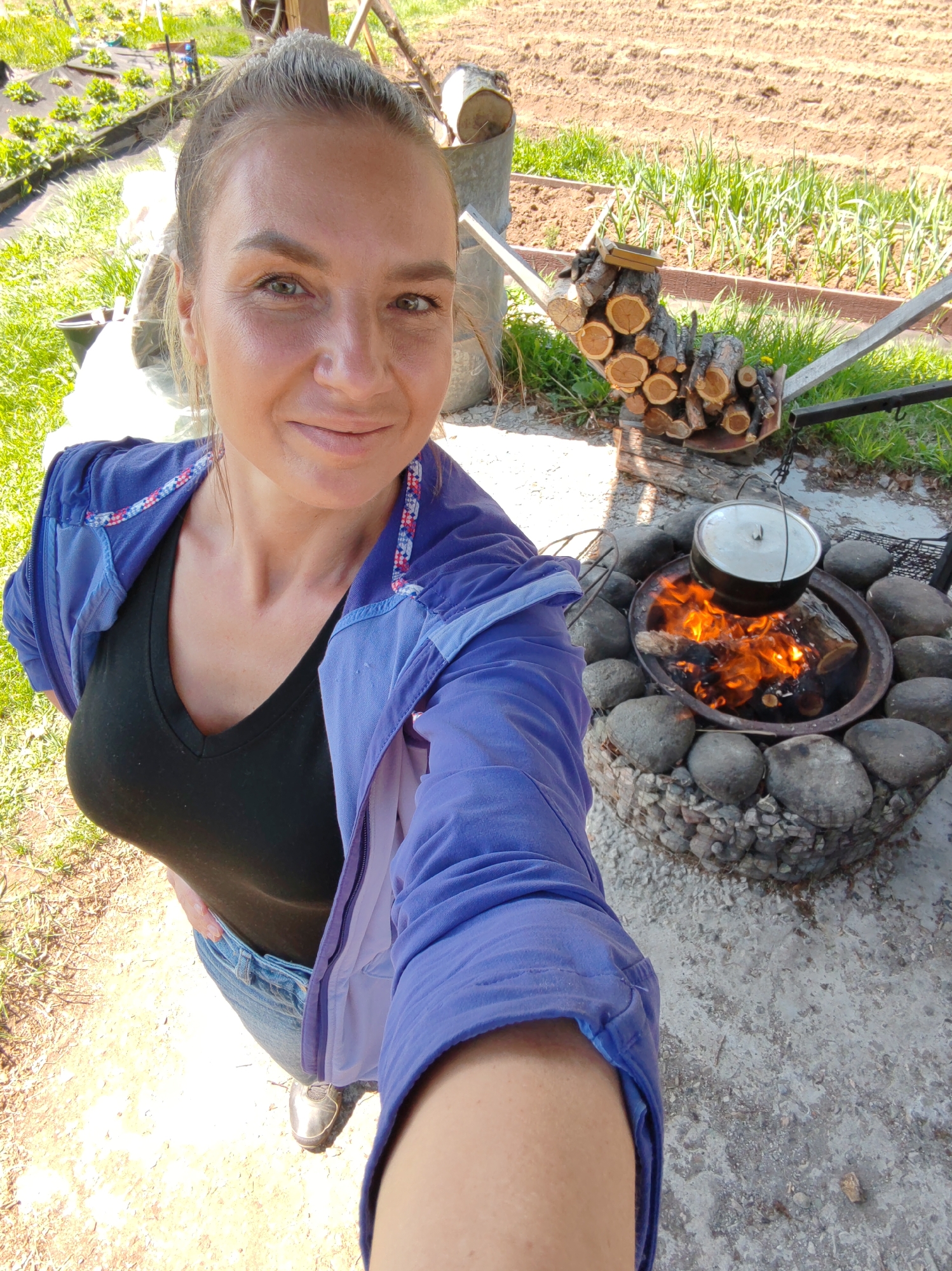 A female subject takes a selfie outdoors in what appears to be a garden or rural setting, location Unknown, Unknown. She directly faces the camera with a slight smile, extending her right arm to hold the device. Her hair is pulled back in a bun, and she exhibits visible facial lines around her eyes. She wears a black V-neck top, blue denim jeans, a vibrant blue zipper jacket with a patterned lining visible at the collar, and metallic-colored sneakers.
To the subject's right and slightly behind, a circular fire pit constructed from large, rounded stones is visible. Within the pit, an active wood fire displays bright orange and yellow flames. A metal pot with a lid hangs suspended by a chain over these flames, attached to a black metal stand with hooks. Adjacent to the fire pit, a horizontal wooden structure or log holder contains neatly stacked, cut logs of various sizes.
The background features multiple garden elements. To the left, a raised bed with small green plants is partially covered by black fabric. Further back and to the right, a large tilled field with distinct rows of bare earth is visible, alongside another garden bed containing taller green shoots, possibly garlic or onions. The ground is an uneven mix of dirt, grass, and a lighter-colored paved or concrete area around the fire pit. The scene is brightly illuminated by direct sunlight, creating strong shadows.