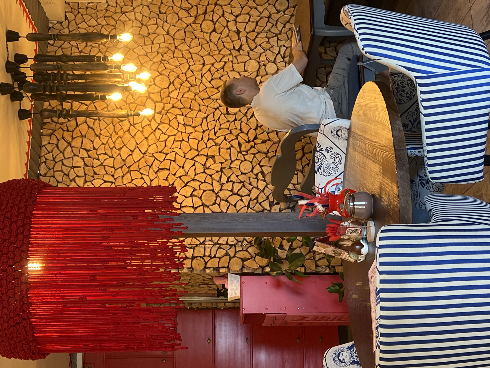 The image depicts the interior of a restaurant in Chelyabinsk, Russia. A man wearing a white t-shirt and grey pants is seated at a table, looking to his right. He appears to be interacting with something on the table, possibly a tablet or a book. The restaurant features a distinctive wall covered in cut logs arranged in a circular pattern, and a large red chandelier with hanging strands. The furniture includes chairs upholstered in blue and white stripes and a chair with a matryoshka doll pattern. On the table, there are menus, a small vase with red and blue feathers, and a sugar bowl. Red cabinetry and a red cabinet are visible in the background. The lighting is warm, with several exposed filament bulbs in a wooden chandelier hanging from the ceiling.