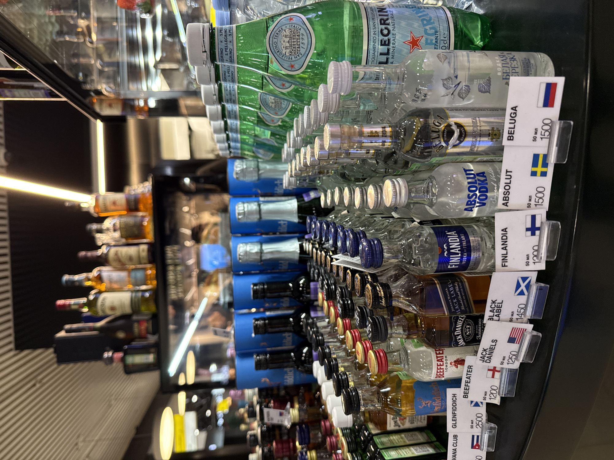 The image displays a retail store counter in Khimki, Russia, featuring a meticulously arranged selection of miniature alcoholic beverages and sparkling water bottles. The foreground is dominated by multiple rows of 50ml "nip" bottles of spirits, including brands such as Havana Club, Glenfiddich, Beefeater, Jack Daniel's, Johnnie Walker Black Label, Finlandia, Absolut Vodka, and Beluga Noble Vodka, along with "Белая березка" vodka. Each miniature bottle is accompanied by a white price label in a clear plastic holder, displaying the brand name, "50 мл" volume, a numeric price (e.g., 1200, 1500, 2500, likely in Rubles), and the national flag corresponding to the liquor's origin.
To the right, a row of approximately eight green glass bottles of San Pellegrino sparkling natural mineral water is prominently displayed, featuring white screw caps and the distinctive green label with a red star. Behind these miniature and sparkling water bottles, larger liquor bottles are visible, some in blue cylindrical packaging (Glenfiddich) and others with silver foil-wrapped necks resembling sparkling wine or champagne. On an elevated shelf in the mid-background, full-sized bottles of various spirits, including Jameson Irish Whiskey and Campari, are displayed. The background is blurred, showing additional retail shelving, a refrigerated display case with internal lighting, and overhead artificial lighting elements, including linear fluorescent lights and spotlights, illuminating the scene. The ceiling above features a light-colored, possibly slatted or ribbed, design. No individuals are present in the image.