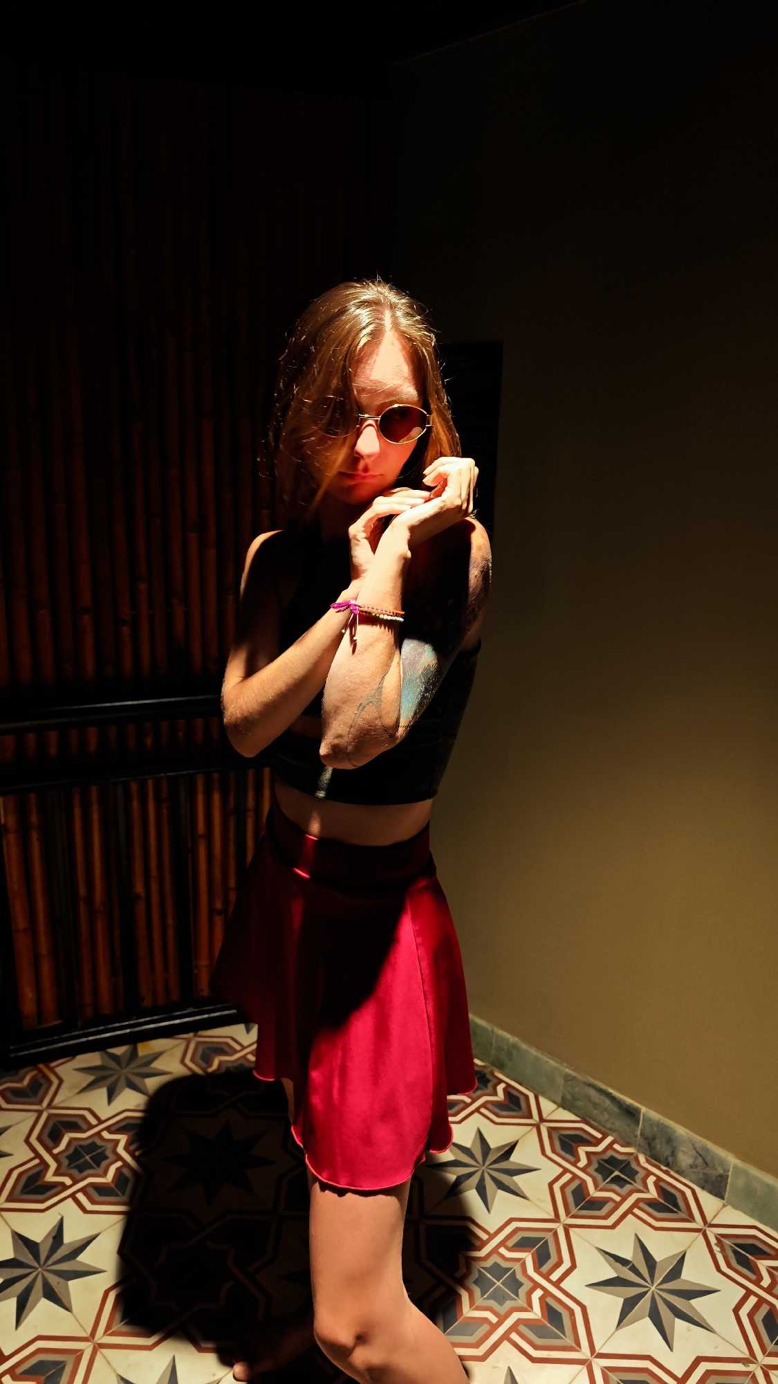 The image captures a single individual, appearing to be a young adult female, posing indoors under dramatic, directional lighting. She has long, light brown hair and wears dark-lensed, oval-shaped sunglasses, a dark crop top, and a bright red short skirt. Her forearms are crossed in front of her, with her hands resting near her shoulders. On her right wrist, a pink string bracelet and a multi-beaded bracelet are visible. Her left upper arm displays a multi-colored tattoo with intricate designs. The individual is barefoot. The background on the left consists of a dark, vertically slatted wooden or bamboo wall with a lower horizontal barrier, largely obscured by shadow. To the right, the background is a plain, dark-toned wall. The floor is covered with ornate geometric tiles featuring star and octagonal patterns in shades of brown, beige, off-white, and dark grey. A distinct, elongated shadow of the person is cast on the tiled floor, consistent with a strong light source positioned to her front-left. This scene is located in the city of Mu Cang Chai, Vietnam.