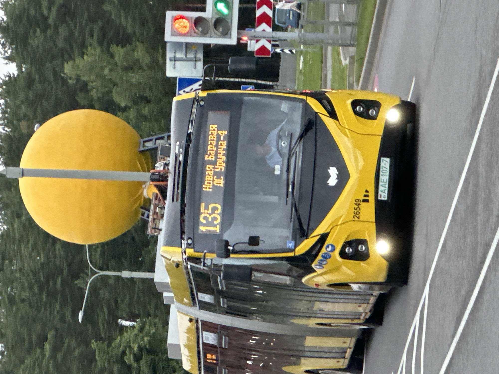 This is a vertically oriented, eye-level, medium shot of a yellow and black bus stopped at a traffic light. The bus is the primary subject, with its headlights on and illuminated. The front of the bus has a digital display showing the number "135" in yellow characters. To the right of the number, text in Cyrillic script is visible, reading "Новая Баравля" and "Дс Уручча-4". A license plate with the characters "ААЕ 107-7" is also visible on the front right side of the bus. To the left of the bus, a large, bright yellow sphere is partially visible, attached to a pole. This sphere appears to be some kind of public art or fixture. In the background, there are trees and traffic lights, with one light showing orange. The scene suggests a public road or street. The weather appears overcast, and the time of day is likely daytime due to the ambient light. The image is slightly blurry, suggesting it was taken while moving or from a distance.