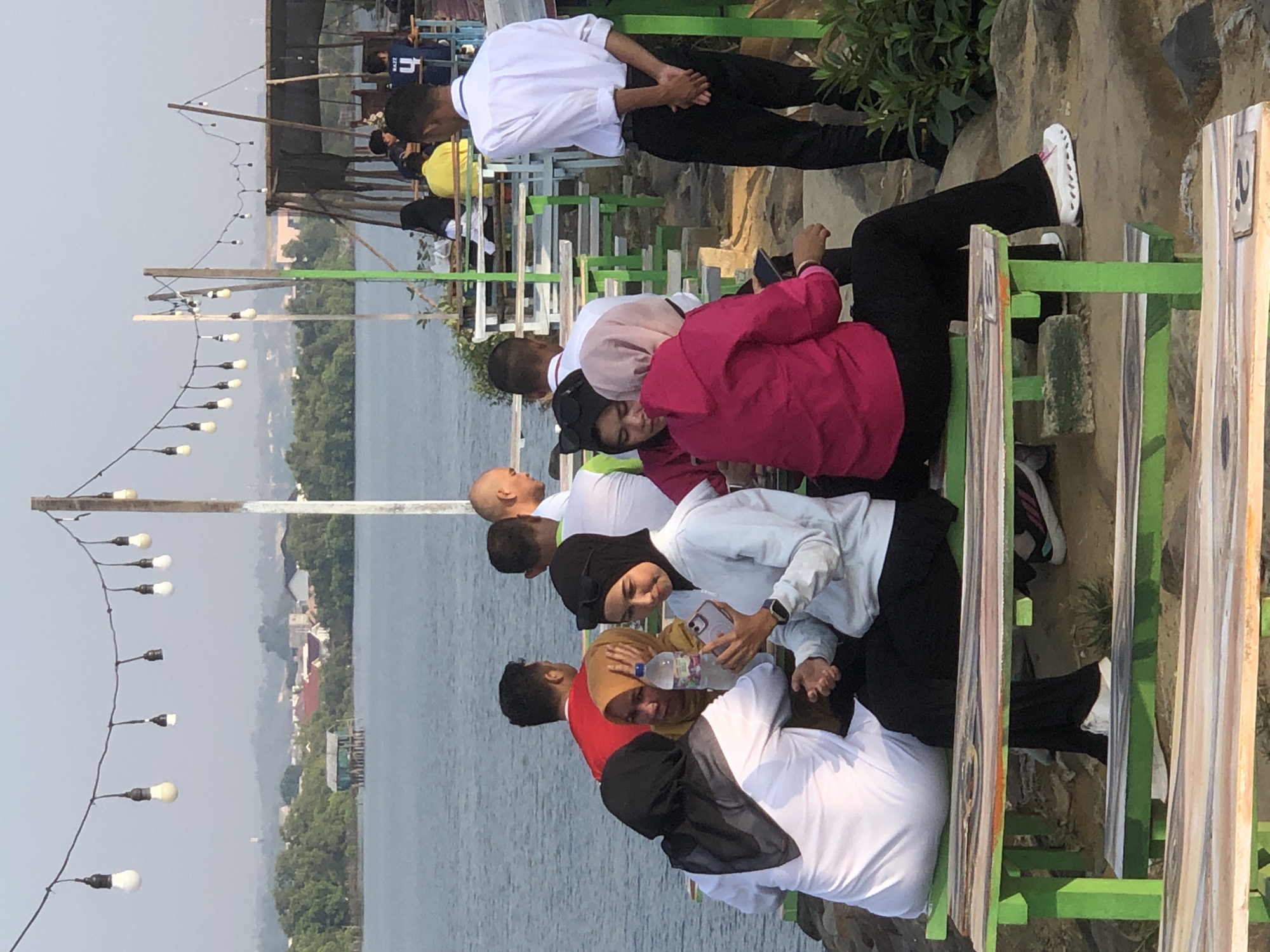 A group of people are gathered at an outdoor cafe by the water in Lhokseumawe, Indonesia. The cafe appears to be set up on a slightly elevated platform overlooking a body of water. String lights hang overhead, and wooden picnic tables with green frames are arranged for seating.
Several individuals are seated at one of the tables, engaged in conversation and using their phones. One woman in a white hijab and a white top is holding a smartphone and looking at it. Another woman next to her wears a pink hooded jacket and black pants. A third person, wearing a yellow hijab, is seated with her hand on her head. A man in a red shirt is also visible at the table.
In the background, there is a view of the water, with trees and buildings on the distant shore, suggesting it's a coastal or lakeside area. Another man in a white collared shirt and black pants stands near the tables, appearing to be part of the cafe staff. The overall atmosphere suggests a casual gathering or dining experience.