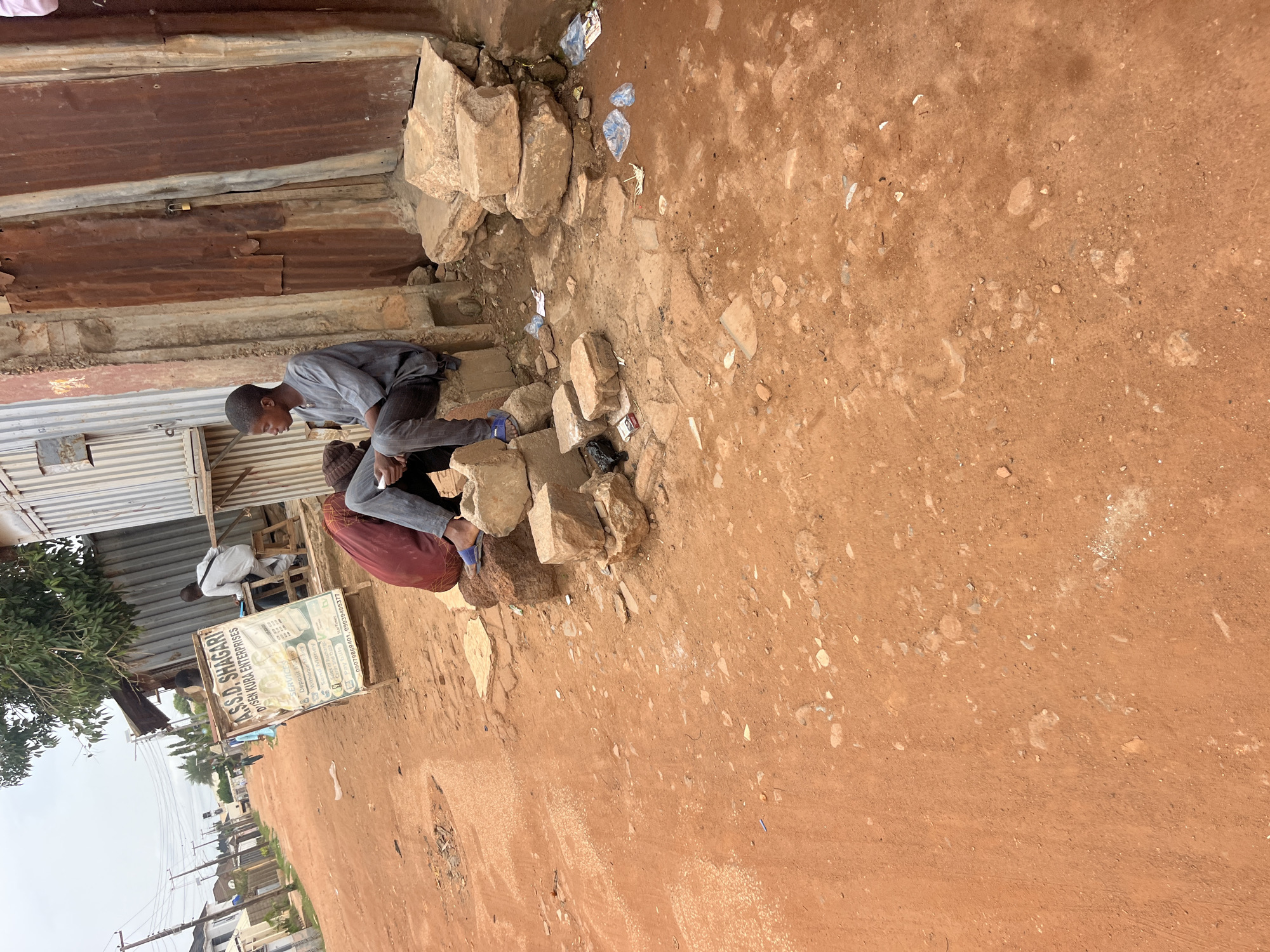 In an outdoor, urban environment with a dirt road, two young men sit on rocks and bricks. One man, wearing a grey shirt and dark pants, sits on a concrete block, holding a white object in his hands and looking to the left. The other man, partially obscured by a burgundy cloth, wears blue slippers. Behind them, a weathered metal structure with corrugated siding and a wooden shelf stands next to a rusty metal door. A sign advertising services like "Send Money" and "Deposit Money" is propped up on an easel. The ground is covered in red dirt, scattered with small stones and litter, including plastic bags and wrappers. In the background, a street lined with houses and utility poles recedes into the distance under an overcast sky.
