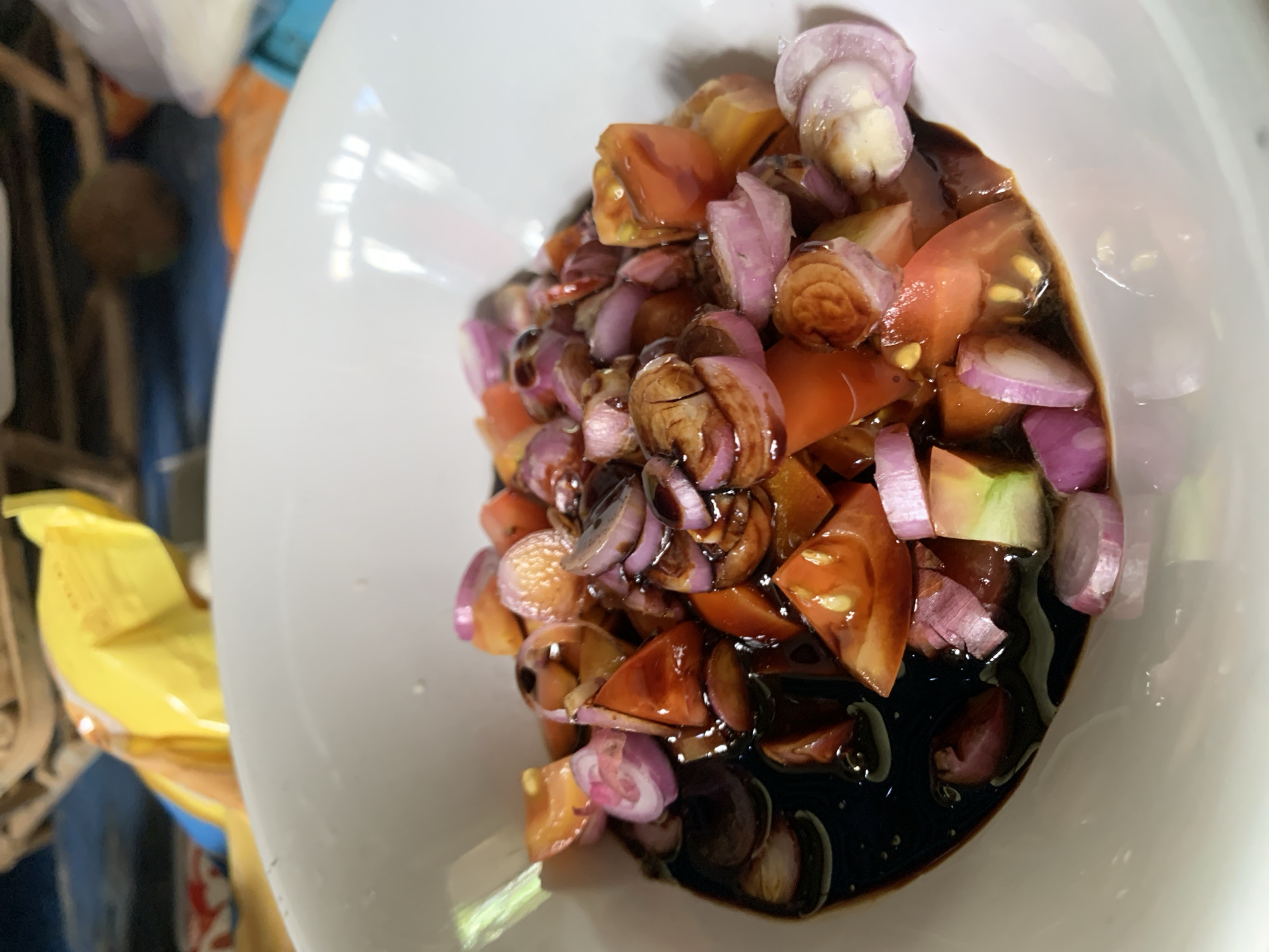 A close-up shot in Karangkemiri, Indonesia, reveals a white bowl filled with a mixture of sliced tomatoes and shallots coated in a dark, glossy sauce, possibly soy sauce or a similar condiment. The bowl's interior is mostly occupied by the tomato and shallot mixture, with some of the dark sauce pooling at the bottom. Part of a yellow package is visible on the left. The context surrounding the bowl suggests a setting where food preparation or serving is taking place.