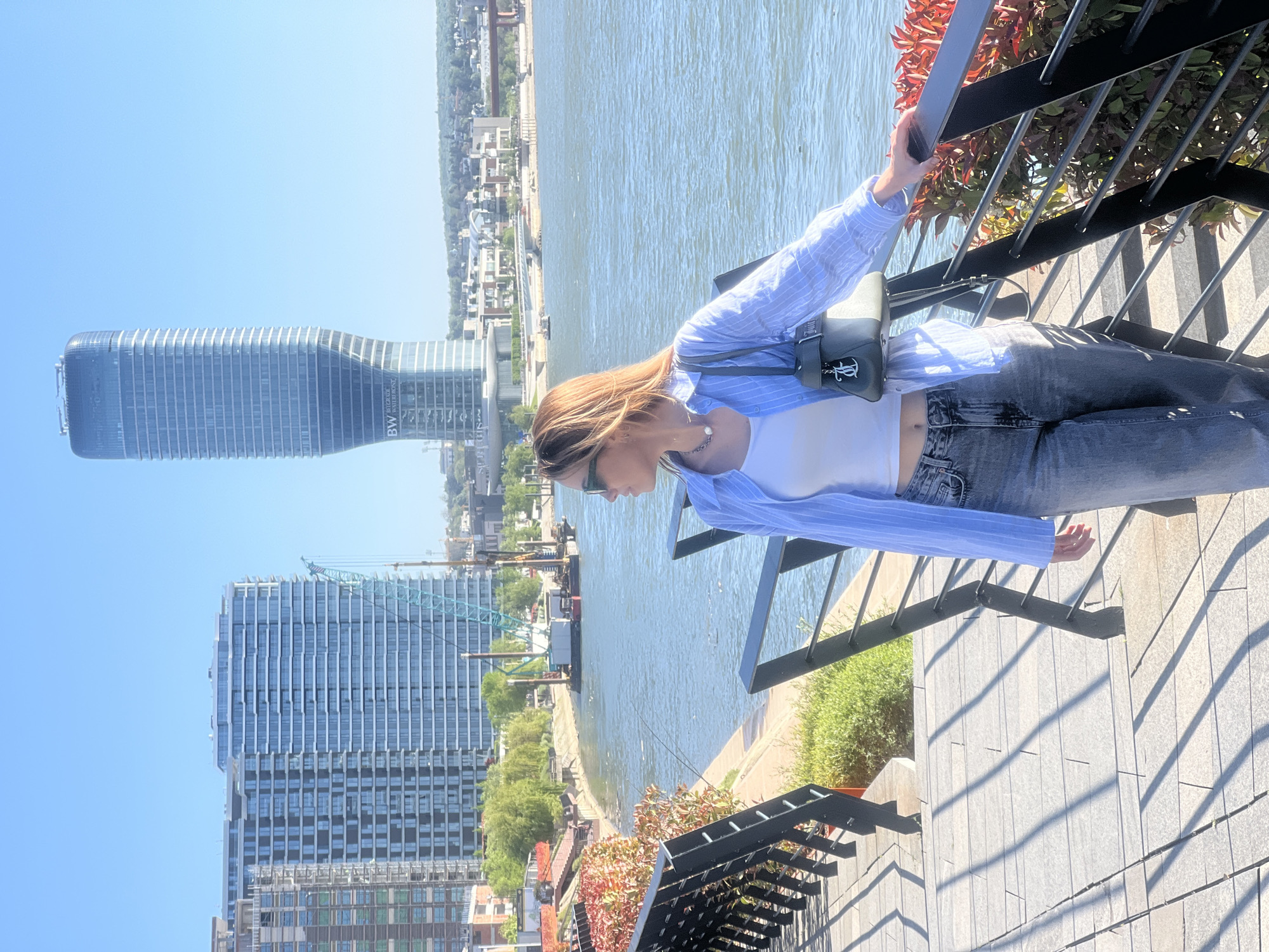 An outdoor, brightly lit image captures a young woman standing on an elevated promenade or terrace, leaning gently on a black metal railing. She is presented in a profile view, looking towards a wide river and a distant city skyline under a clear, sunny sky. The woman has light brown hair and wears sunglasses, a white crop top, an open light blue and white striped button-down shirt, and dark-wash jeans. A small, dark shoulder bag with a metallic emblem hangs across her body.
In the background, a large river flows, bordered by a modern urban landscape. A prominent, tall, and distinctively curved glass skyscraper dominates the left side of the frame (when viewed upright), clearly displaying the text "BW BELGRADE WATERFRONT" on its lower section, indicating Kula Belgrade. Another large, multi-story building, appearing modern and possibly part of the same development, is visible further left, with partial "BW" text also seen. The scene is bathed in bright daylight, casting crisp shadows from the railing onto the light-colored tiled ground. Red and green foliage adds natural elements near the railing. The setting is clearly a sunny day in Belgrade, Serbia, specifically in the Belgrade Waterfront area.