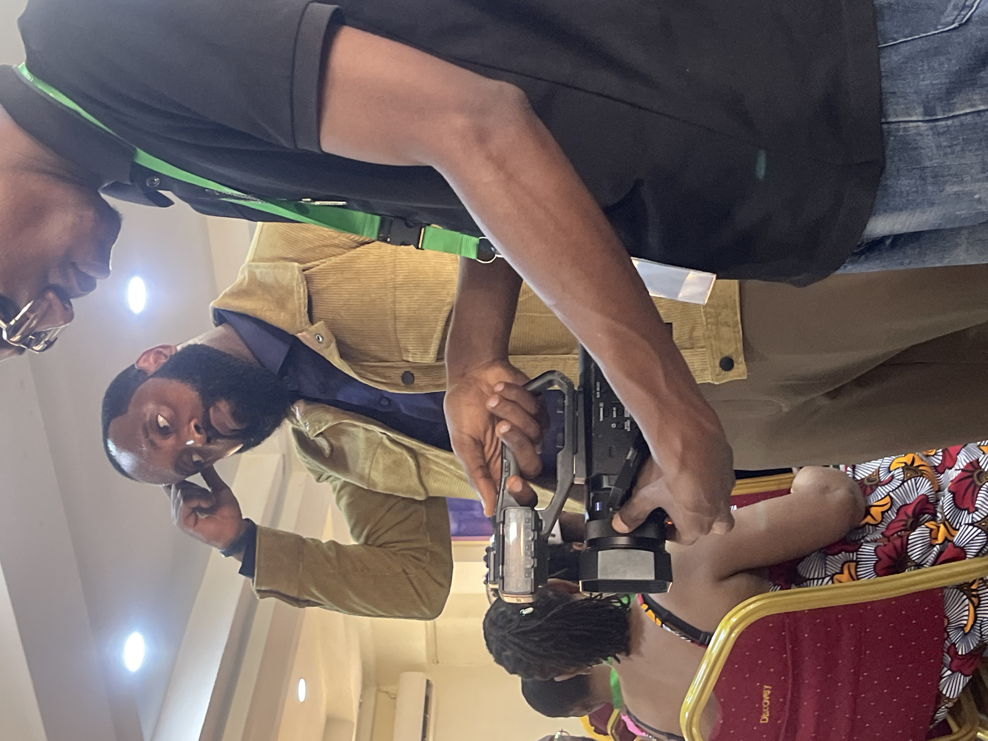The image shows an indoor scene, presumably in Galadimawa Village, Nigeria. Two African men are present. One man is in focus, wearing a beige corduroy jacket over a blue shirt and tan pants. He has a beard and is holding a professional video camera. His finger is touching his chin as if in thought. Another man stands behind and to the side of him, partially visible, wearing a black shirt and jeans, with a green lanyard around his neck and glasses. The camera is pointed towards a person wearing a colorful traditional African print outfit and black dreadlocks, seated on a red chair with a gold frame. The chair has 'Discovery' written on it. An air conditioning unit is visible on the wall in the background. The room is brightly lit by overhead lights.