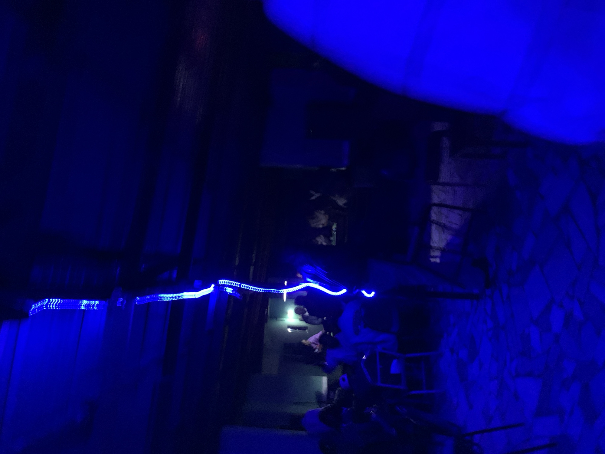 The image depicts an indoor scene within a facility in New Stadium, Nigeria, illuminated predominantly by a strong blue light. A prominent, vertical, winding blue light strip, composed of individual light elements, descends from the ceiling in the central foreground.
Several individuals are present, distributed across the mid-ground and background. Many are seated at tables and chairs, while others appear to be standing. Their specific features and actions are largely obscured by the low light conditions and blue illumination. One person is clearly standing near the base of the central blue light strip.
The environment suggests a social gathering space, possibly a bar or lounge. The floor consists of irregularly shaped, light-colored tiles, which appear unevenly laid. Structural elements, including walls and potentially ceiling beams, are visible but indistinct due to the darkness. A large, out-of-focus blue object occupies the extreme right foreground.