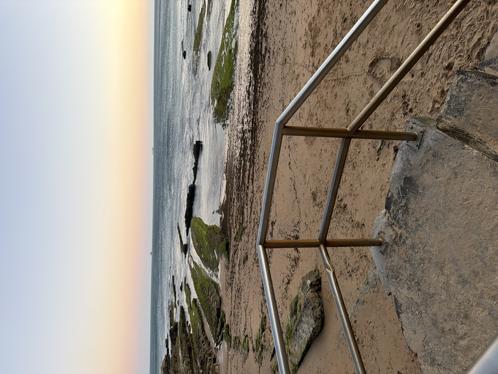 The image captures a sunset view of the coast in Cascais, Portugal. The foreground features a concrete step with a metal railing, leading down to a sandy beach. The beach is scattered with seaweed and rocks, some of which are covered in green algae. The sea stretches out to the horizon, with small waves gently lapping against the shore. In the distance, a few ships are visible on the water, and the sky displays a gradient of pastel colors, transitioning from pale blue at the top to a soft yellow and pink near the horizon. The overall scene conveys a serene and tranquil atmosphere of a coastal evening.