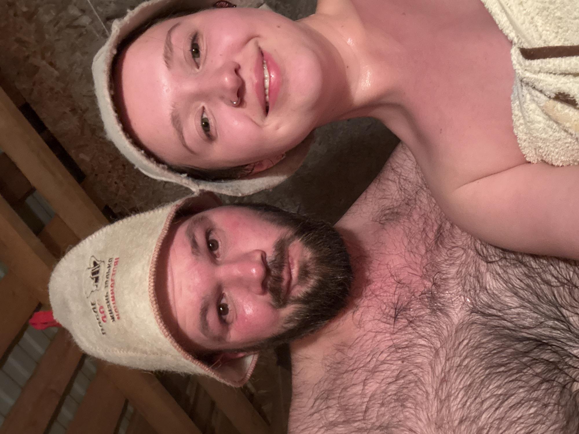 A selfie image features two individuals, a woman and a man, likely inside a sauna. Both are wearing felt hats. The woman, positioned at the top of the frame, is smiling, and her skin appears flushed. A towel is draped over her shoulder. The man, below her, has a beard and a noticeably hairy chest. Their skin also appears flushed. The background reveals a wooden structure, which supports the sauna environment. The location is not discernible as being in the city of Русский Вожой, Russia.