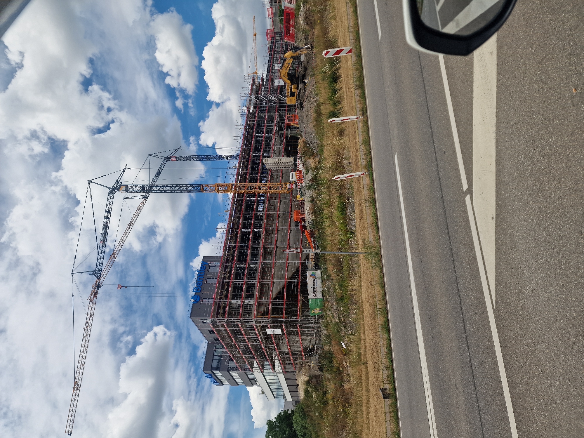 The image shows a construction site in Hechingen, Germany, under a partly cloudy sky. A large, multi-story building, partially covered in scaffolding, is the central focus. The building has "Bentley" branding visible on its facade. Multiple construction cranes are erected around the building, with one large crane extending high above the structure. In the foreground, a road with white lane markings runs across the bottom of the frame. To the right, a car's side mirror is partially visible, reflecting the road and some of the construction site. The surrounding area appears to be a grassy embankment leading up to the construction zone. A yellow excavator is visible on the construction site.