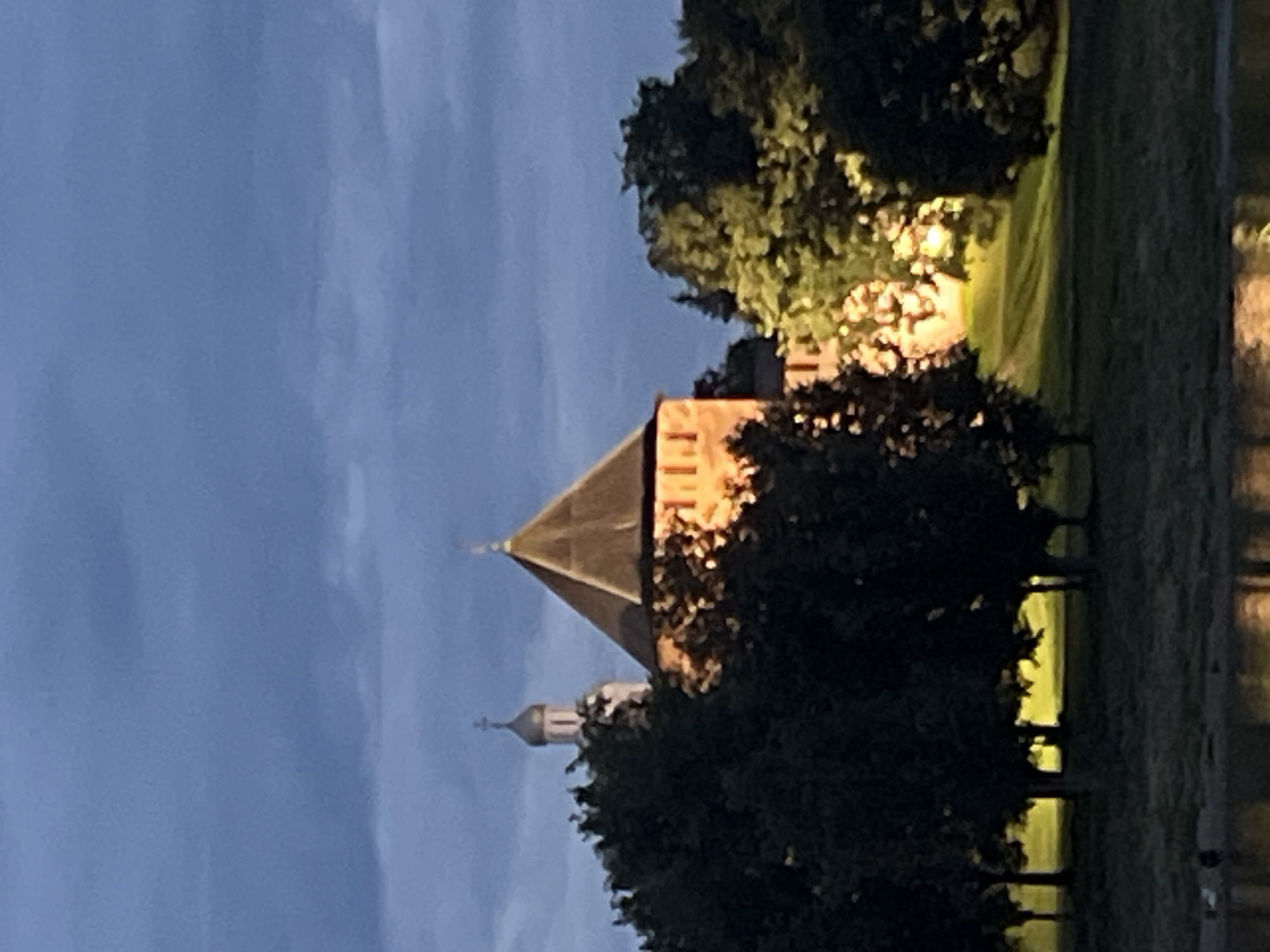 The image shows a twilight view of the Novgorod Kremlin in Veliky Novgorod, Russia. In the foreground, a body of water reflects the light. The middle ground is occupied by trees. Beyond the trees, a brick tower with a conical roof is illuminated. To the left of the tower, a white church with a golden dome is visible. The sky is dark blue with clouds.