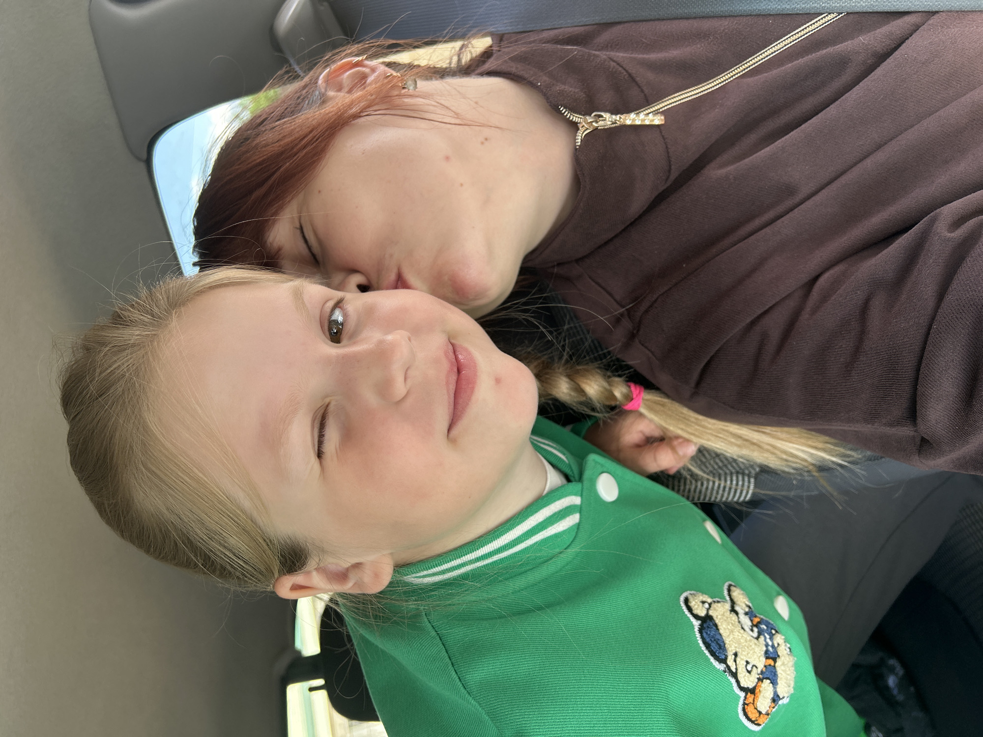 A fair-skinned adult female with auburn hair and wearing a brown top kisses a fair-skinned child with blonde hair, who is wearing a green jacket and winking, on the cheek. They appear to be inside a vehicle, given the car interior and exterior view visible through the window. There is no information about a city or location in Фоки, Russia, visible in the picture.
