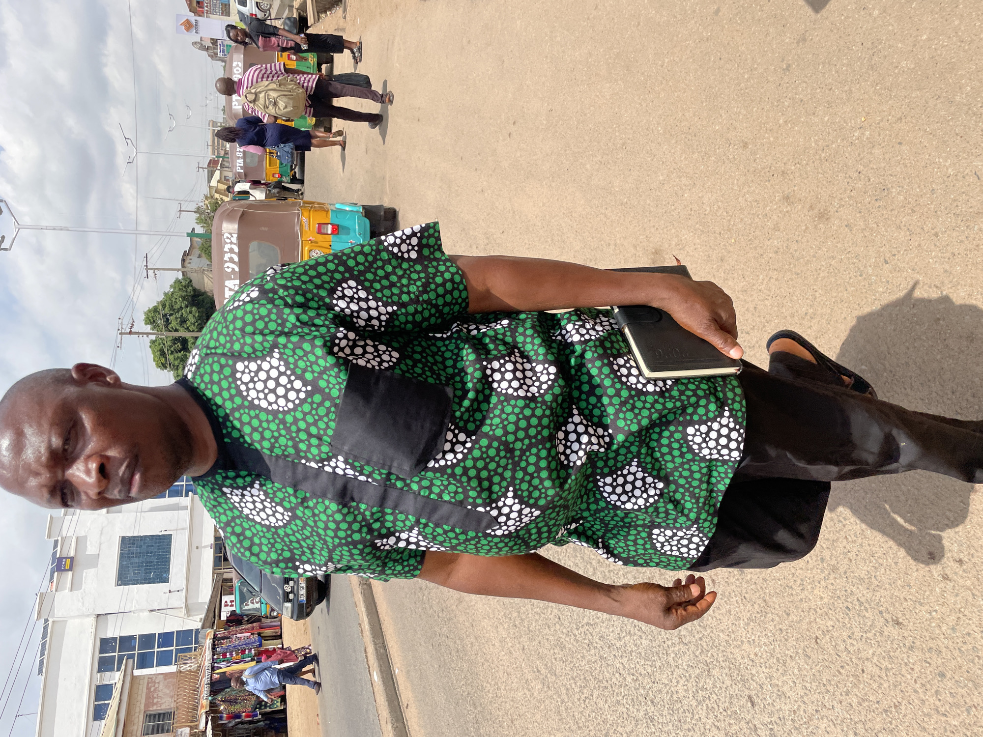 A man stands on a paved street in Jos, Nigeria. He wears a green, black, and white patterned shirt with black trousers and sandals. He is holding a black book. In the background, there are people, commercial tricycles ("keke napeps"), and buildings, suggesting a busy urban environment. The weather appears sunny.