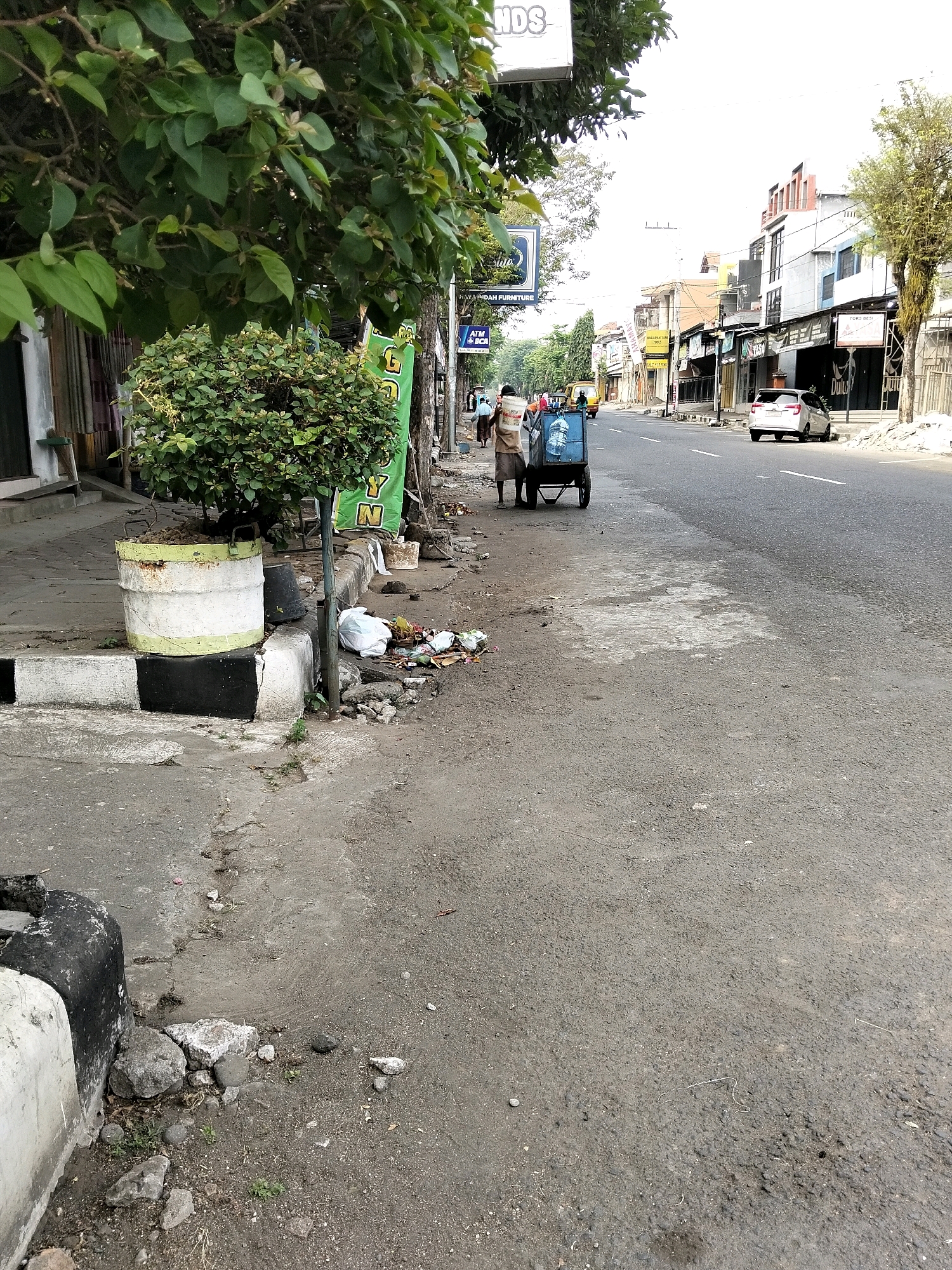 The image depicts a street scene in Purworejo, Indonesia. On the left, a sidewalk is lined with a large planter containing a bush, next to a curb with black and white markings. Beyond the planter, several white plastic bags and scattered debris are visible on the ground. A person is pushing a two-wheeled cart carrying large water jugs. Further down the street, more people are walking, and a white car is parked on the right side. The street is paved, with shops and buildings lining both sides. Trees with green leaves are present along the sidewalk. The sky is bright, suggesting daylight.