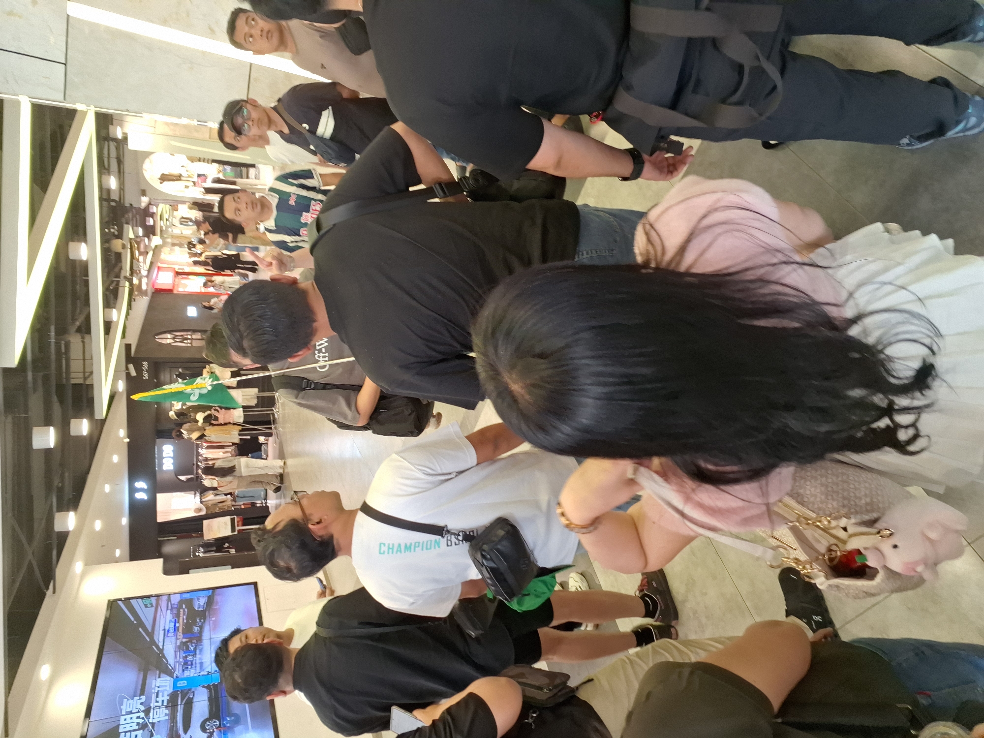 In the Hongkou District of China, a group of people are gathered inside a brightly lit commercial space, possibly a shopping mall. The scene is captured from a slightly elevated, rear-facing perspective, focusing on the backs of several individuals. A large digital screen displays an advertisement, likely for a parking service, featuring an image of a white car and Chinese text. In the background, shops are visible, with mannequins displaying clothing. The individuals in the foreground are dressed in casual attire, with one person wearing a white t-shirt with "CHAMPION" printed on the back, and another in a dark t-shirt and jeans. A person in the front is wearing a light-colored skirt and a pink top, holding a purse adorned with a small pink plush toy. The flooring appears to be tiled.