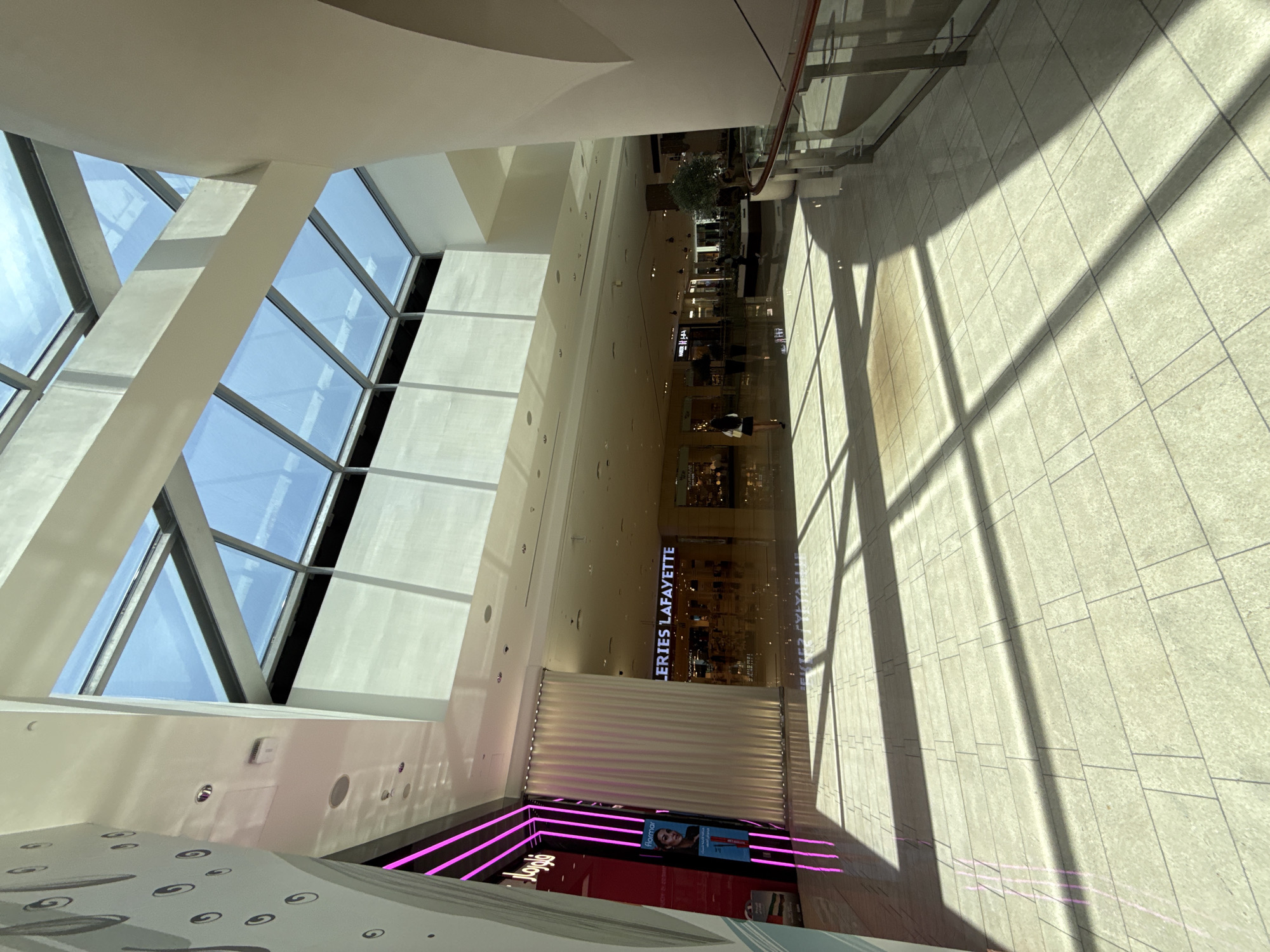 The image shows the interior of a large, modern shopping mall in Dubai, United Arab Emirates. The scene is brightly lit, with natural light filtering through a large glass ceiling structure. The floor is tiled with light-colored square tiles, reflecting the light and creating sharp shadows from the architectural elements.
On the left side of the image, a store with bright pink neon lighting is visible, featuring a display with an image of a woman and text in Arabic and English. Next to it is a tall, floor-to-ceiling curtain. Further into the mall, a store with a prominent sign reading "LERIES LAFAYETTE" is visible. The mall interior is expansive, with multiple levels and walkways.
In the mid-ground, several people can be seen walking through the mall, including a woman in a white top and black skirt walking away from the camera. Shops and displays line the walkways, with some featuring decorative plants.
The architecture is characterized by smooth, curved walls and prominent structural beams supporting the glass ceiling. A modern glass railing with a dark handrail is visible on the right side of the frame, indicating a change in elevation or a walkway overlooking a lower level. The overall impression is one of a clean, spacious, and upscale shopping environment.