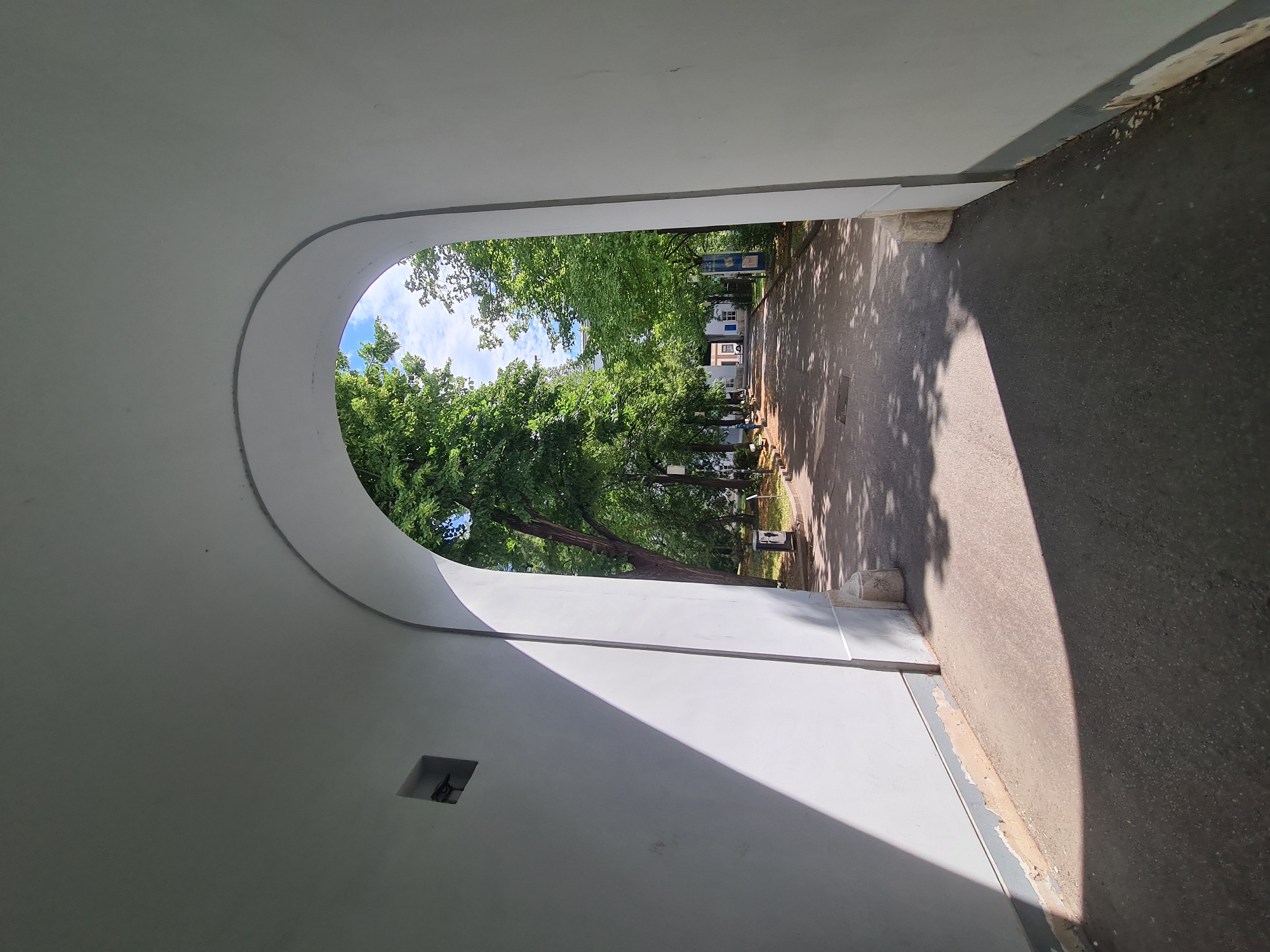 The image shows a sun-dappled pathway leading away from an arched entryway. The entryway is framed by white walls, with a prominent shadow cast across the ground from the left. Beyond the arch, a paved path is lined with mature trees on both sides, casting dappled shadows on the ground. In the distance, buildings with white facades and blue-tiled roofs are visible, suggesting a campus or institutional setting. The sky is blue with scattered white clouds. The scene appears to be in a park or university grounds. This location is the Schönbrunn Palace grounds in Vienna, Austria, specifically the entrance to the gardens from the Gloriette alley.