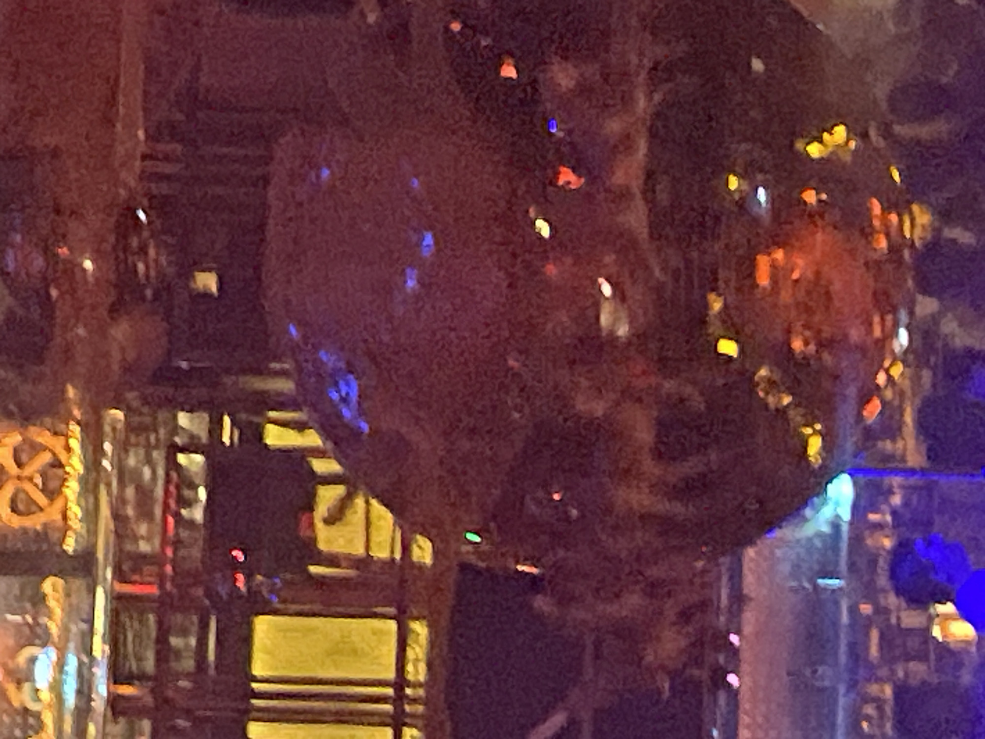 An underexposed, blurry photograph captures an indoor scene, likely a club or entertainment venue, located in the city of Unknown, Unknown. A large, dark, spherical object, possibly a disco ball or highly reflective sculpture, occupies the central and right portions of the frame, reflecting numerous fragmented, multi-colored lights in shades of blue, red, orange, yellow, green, pink, and white. Above the sphere, indistinct architectural elements, possibly metal beams or railings, are visible, with bright yellow light sources positioned behind them. In the upper center, a blurry, illuminated sign resembling a pretzel or an intertwined symbol is present. The lower left foreground shows the head and shoulders of a person silhouetted against a cool blue light. Numerous other blurry figures are dimly discernible in the background and as reflections on the sphere, indicating a gathering of people within the dimly lit environment.