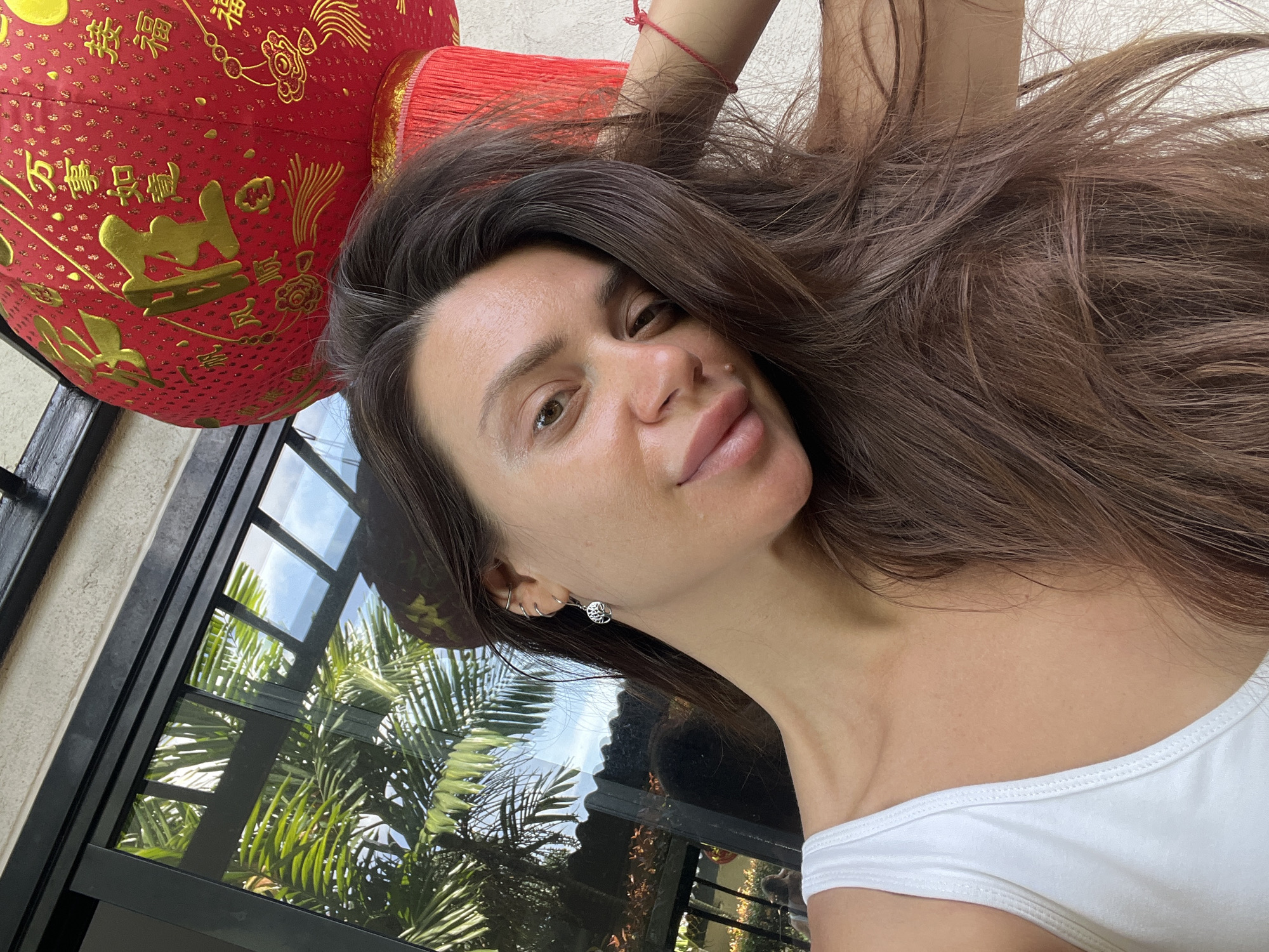 A selfie of a woman with long brown hair wearing a white top, next to a red Chinese lantern with gold lettering. The background includes a window with a view of palm trees. Location is Unknown, Unknown.