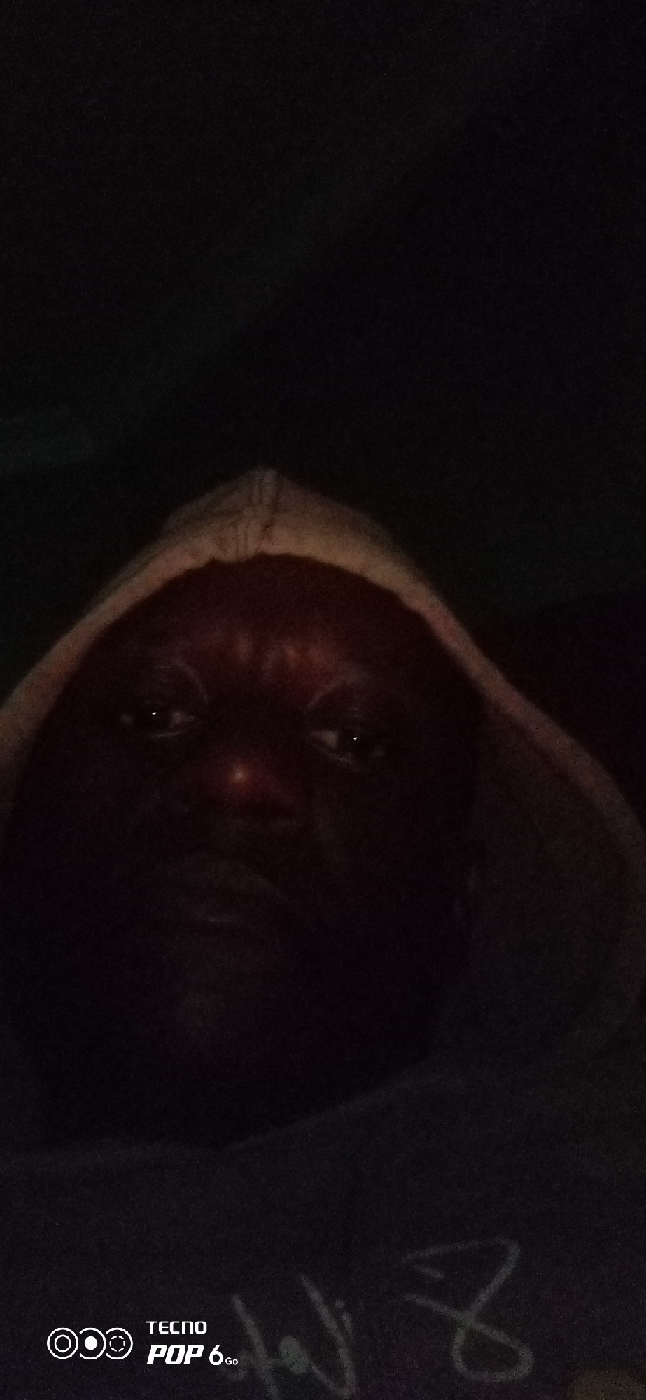A close-up, low-light, selfie-style photo shows a person's face partially obscured by a hood. The person has dark skin and is looking directly at the camera. The image is dark and grainy, with the hood creating shadows that obscure much of the face. In the bottom left corner, there is a logo that reads "TECNO POP 6 Go." The background is a dark, indistinct mass. There are no specific geographical markers to indicate the location in Jos, Nigeria.